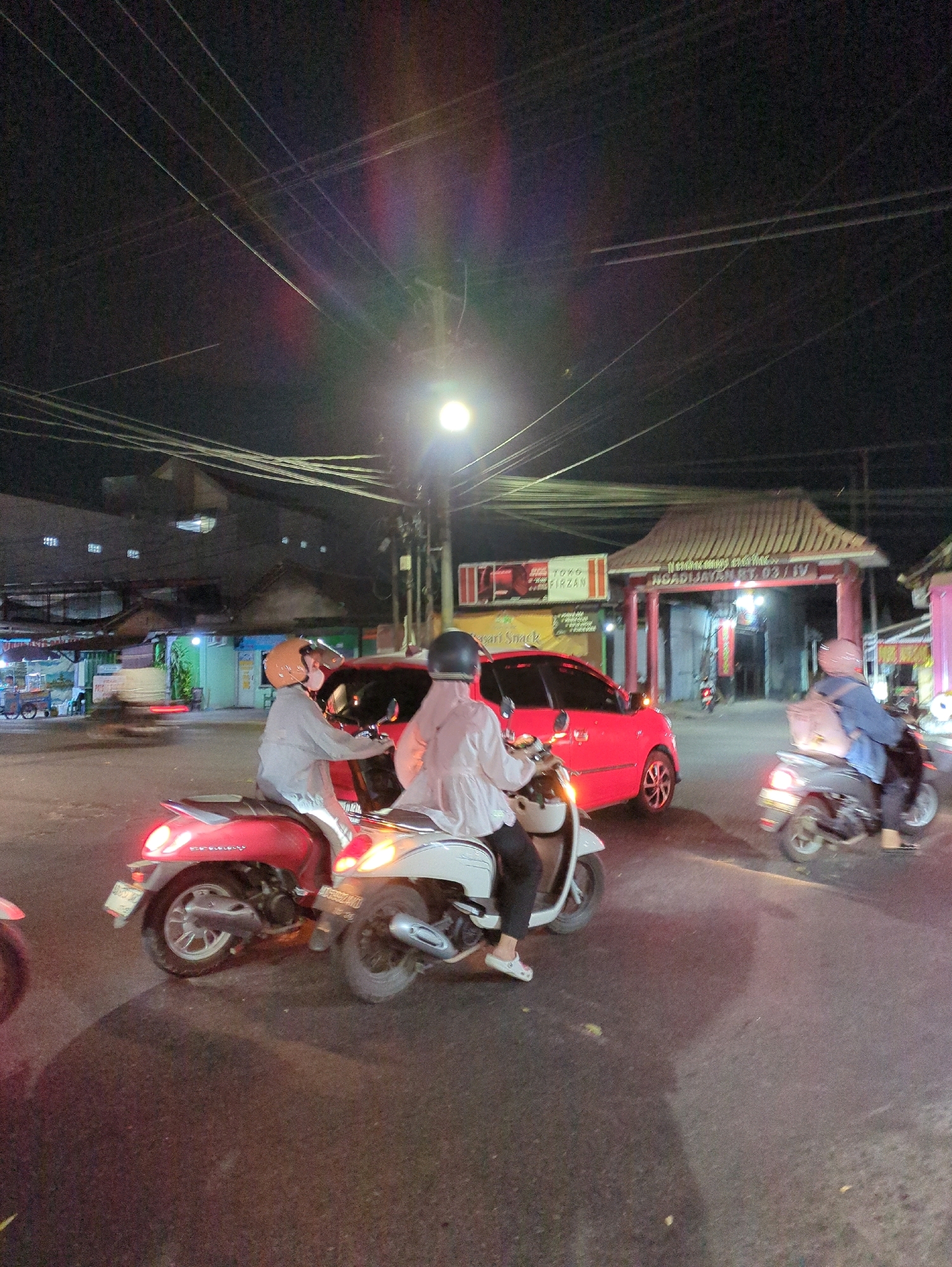 A nighttime street scene in Gumpang, Indonesia, shows several motorbikes and a red car at an intersection. Two people on a red motorbike are in the foreground, with one driver and a passenger wearing helmets. To their right, two more individuals on a white motorbike are also visible, with the rider wearing a helmet and a passenger in the back with a light-colored headscarf. A red car is stopped behind the red motorbike, partially obscuring a building entrance with a traditional roof structure. Another motorbike with a rider in a blue jacket and light-colored headscarf is moving away from the camera to the right. Overhead, a dense network of electrical wires crisscrosses the dark sky, illuminated by a street light and the headlights of the vehicles. The asphalt road surface shows signs of wear and tear.