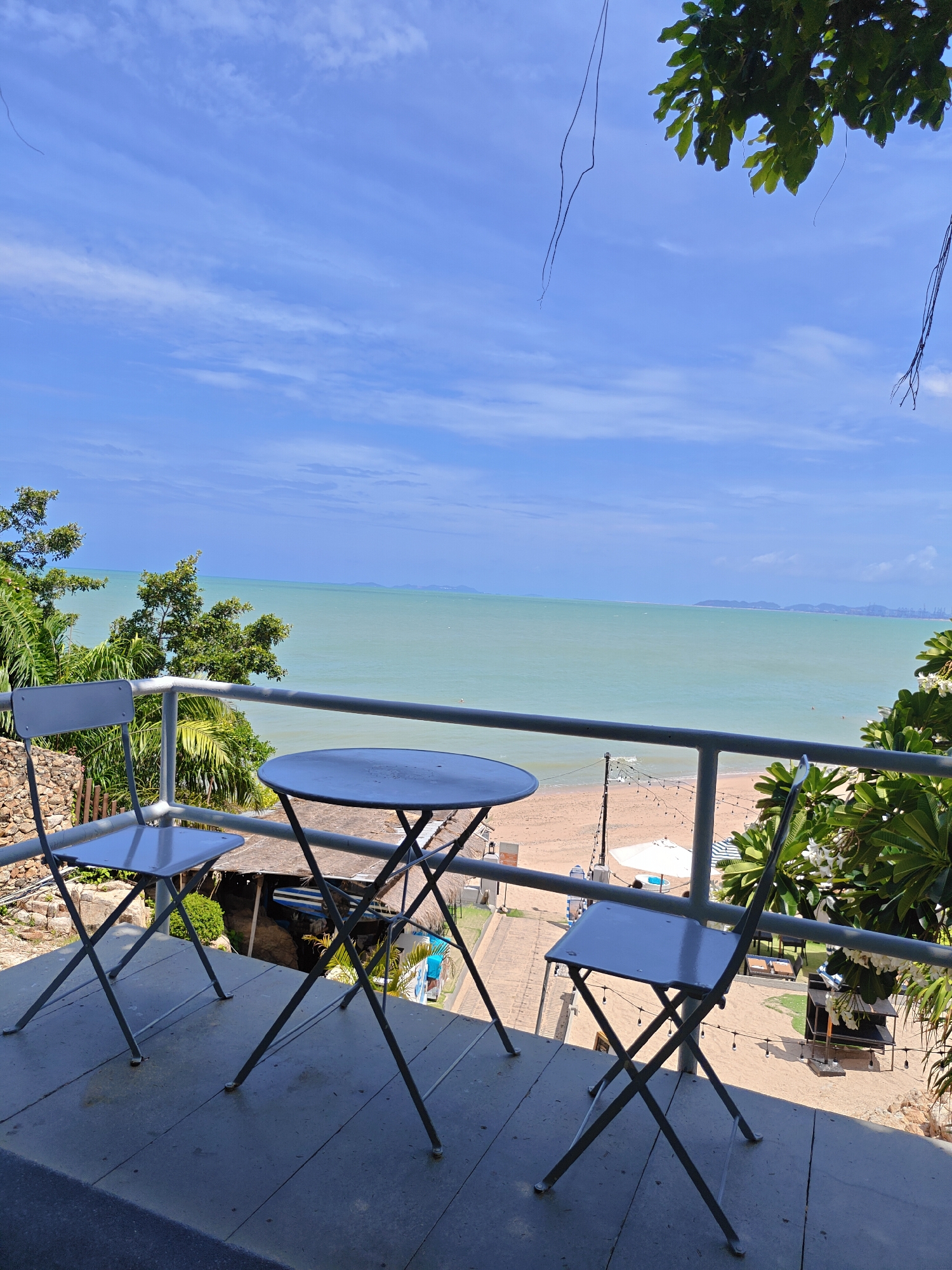 The image captures a picturesque view from a balcony overlooking the ocean in Pattaya City, Thailand. In the foreground, a round metal table with two matching metal chairs are arranged on a wooden deck, suggesting a seating area for a cafe or restaurant. The chairs and table are gray and have a simple, foldable design. Beyond the balcony, a sandy beach stretches out, dotted with a few white umbrellas and lounge chairs. The turquoise ocean water is calm, with a faint haze visible on the horizon where landmasses can be seen. Lush green trees and tropical foliage surround the balcony, adding to the serene atmosphere. The sky is a vibrant blue, with scattered white clouds. The overall scene conveys a sense of relaxation and tranquility, typical of a seaside resort.