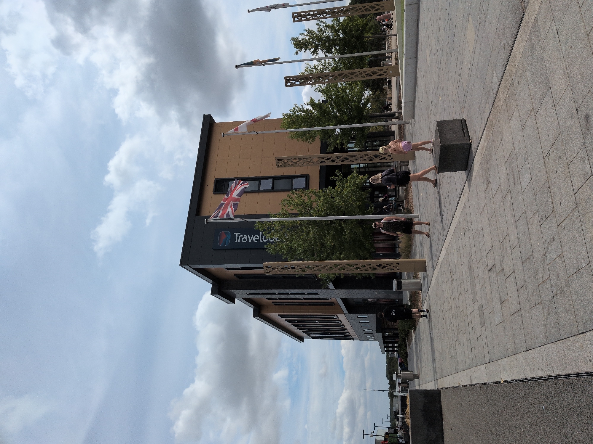 The image captures a modern building, likely a Travelodge hotel, situated in Telford, United Kingdom. The building features a dark grey facade with lighter brown panels and large, dark-framed windows. A prominent "Travelodge" sign is visible on the side of the building. Several flags are displayed on flagpoles in front of the hotel, including the Union Jack and others that are not clearly identifiable.
In the foreground, a paved walkway is lined with decorative metal structures and trees. The walkway is populated by several individuals: a person in a black hoodie walks away from the camera, a woman in a black top and shorts walks towards the camera, and a young girl in a pink top and shorts walks in the same direction. Further in the background, other people are visible, some sitting on benches and others walking. The environment suggests a commercial or hospitality area, with a cloudy sky overhead.