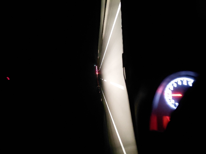 A nighttime view from inside a car shows the road ahead with glowing white lines. Tail lights of a distant vehicle are visible. The dashboard speedometer, illuminated with white numbers and a red needle, is partially obscured by a dark silhouette. The scene is predominantly dark.