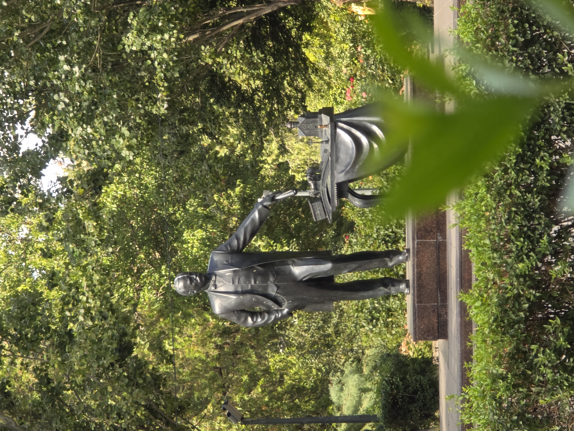 This is a medium shot photograph of a bronze statue of a man in a suit standing in a park setting in Rostov-on-Don, Russia. The statue depicts a man with a mustache, wearing a suit, vest, and bow tie, with his left hand in his pocket and his right hand extended towards a table. On the table, there are miniature architectural models, possibly of buildings, and what appears to be a lamp. The statue is on a pedestal made of stone. The background is filled with lush green trees and foliage, with some red flowers visible in the middle ground. The lighting suggests it is daytime, with sunlight filtering through the trees. In the foreground, there is a blurry green bush, partially obscuring the lower portion of the statue and the surrounding greenery.