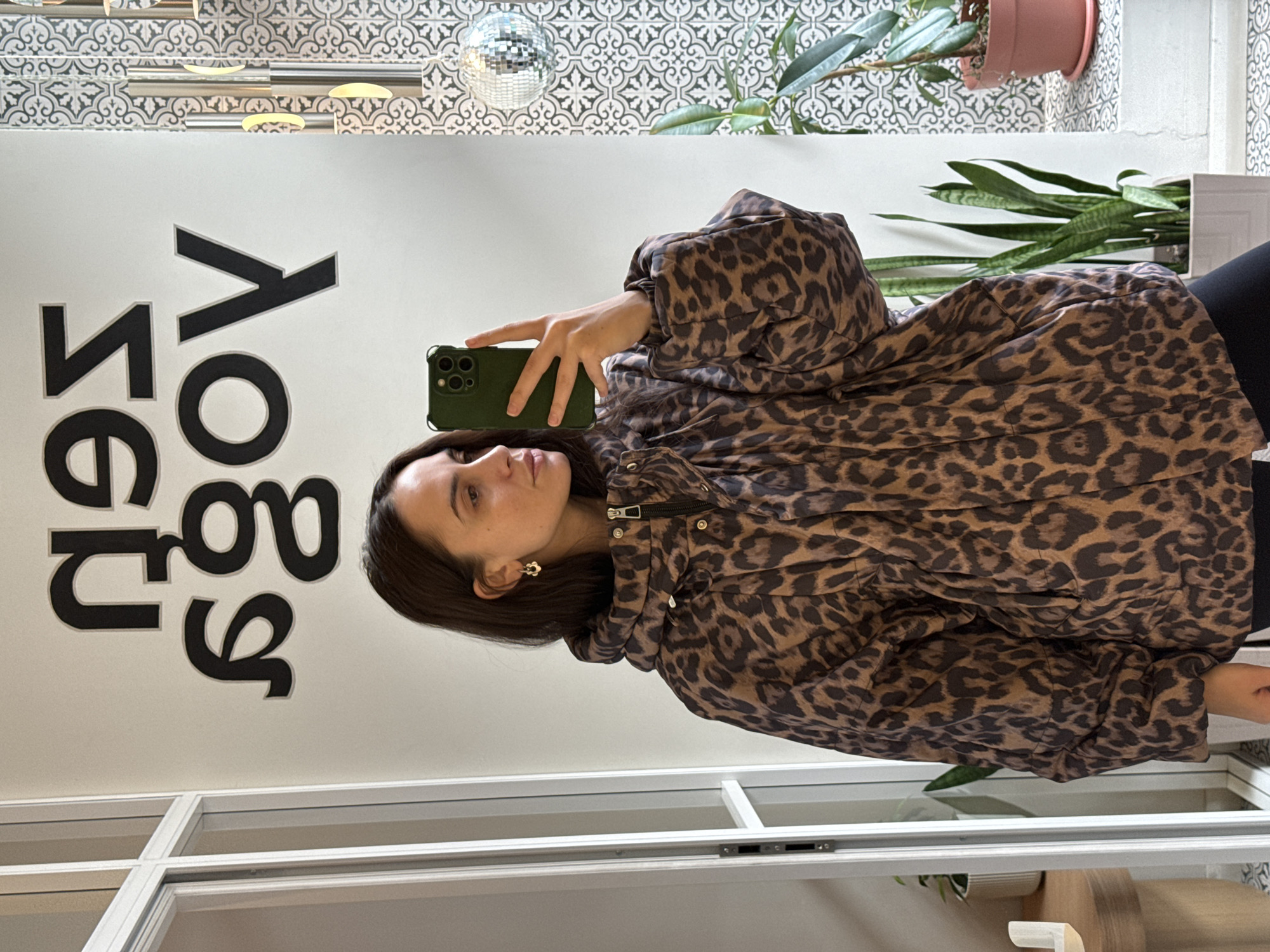 A woman with dark hair and light skin is taking a selfie with a green smartphone in a brightly lit interior space. She is wearing a brown and black leopard-print jacket. Her left ear has a flower-shaped earring. Behind her is a white wall with vertically aligned text that reads "Yoga Segezha" in black lettering. Above the text and woman are decorative patterned tiles and a disco ball hanging from the ceiling. In the background, to the right, are plants in pots and a white planter box. White framed windows or partitions are visible in the foreground and lower part of the frame. The location is Krasnoyarsk, Russia, as indicated by the "Yoga Segezha" text, which references a Segezha Group yoga studio location.