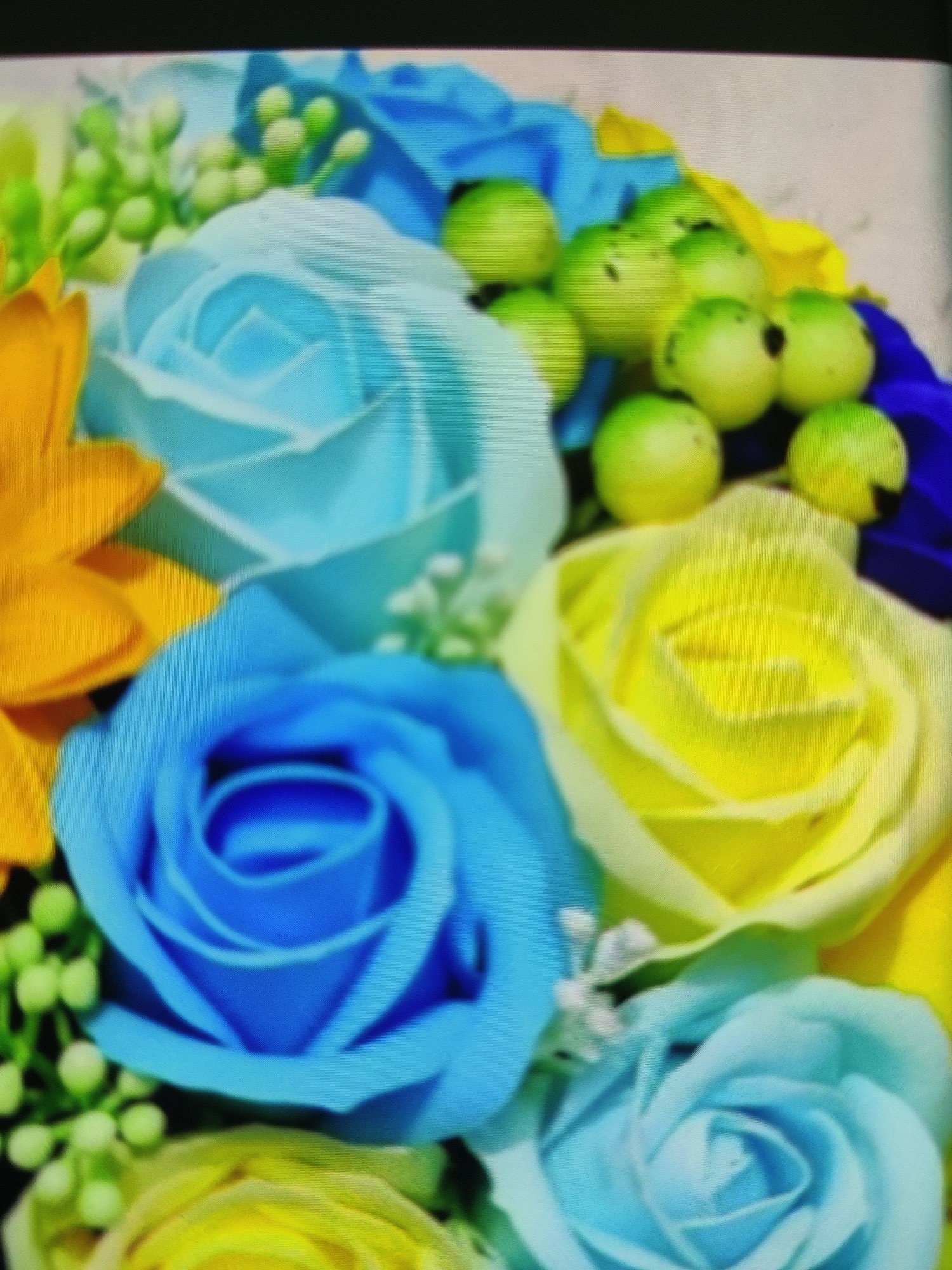 A close-up, overhead shot shows a bouquet of artificial flowers. The bouquet contains several large roses in shades of bright blue and yellow. Smaller, green floral accents with tiny buds are scattered throughout. Some dark blue and yellow fabric is visible behind the flowers. The overall impression is of a vibrant and colorful arrangement. The image was taken in Lower Paxton Township, United States of America.