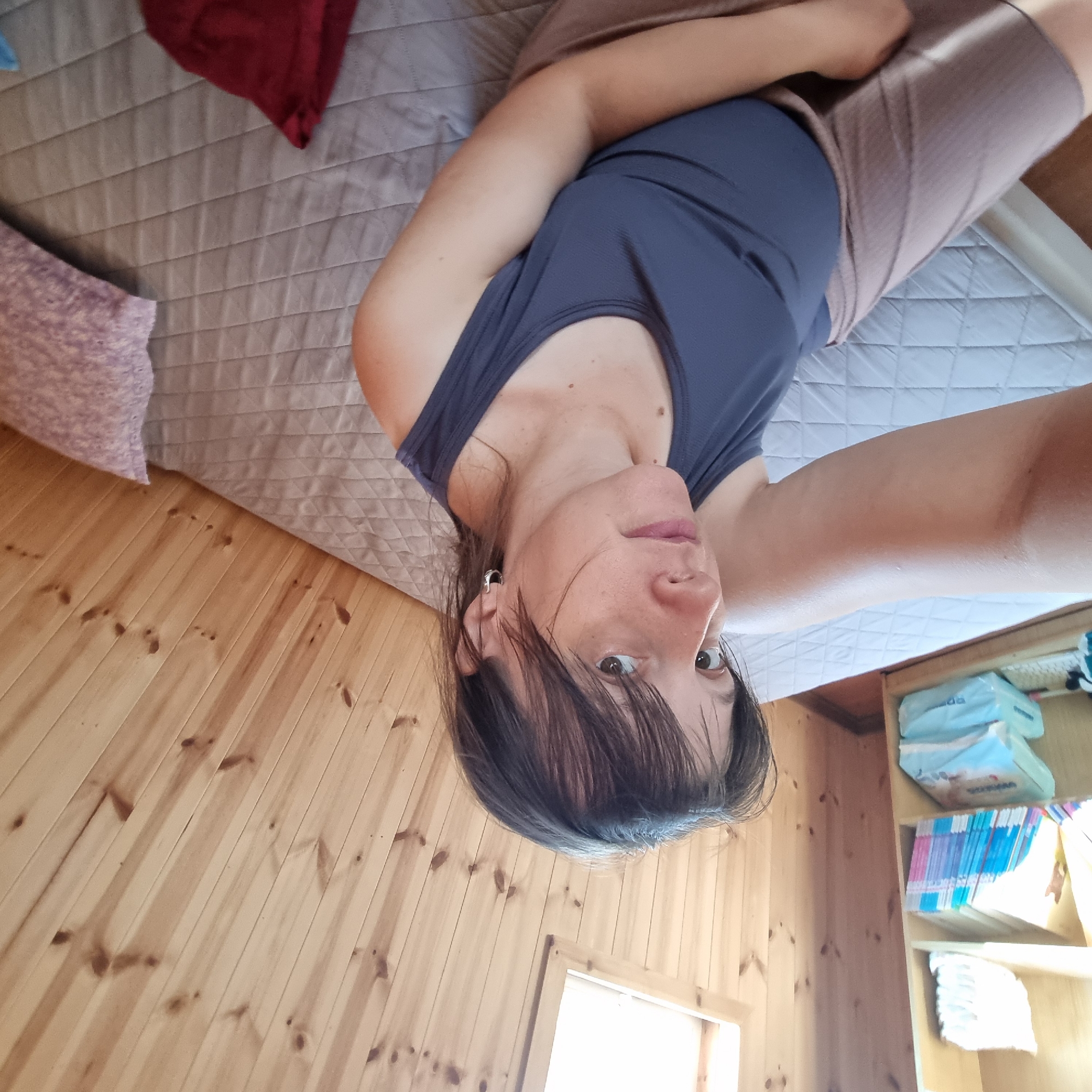A woman with shoulder-length brown hair and fair skin is lying on a bed in a room with wood-paneled walls and ceiling. She is wearing a dark blue tank top and a light brown skirt. Her left arm is extended towards the camera. To her left, a bookshelf filled with books and packages is visible. The bed has a gray quilted mattress and a light purple pillow. A red cloth is also on the bed. The scene appears to be indoors, possibly a bedroom or a living space in a residential setting. There are no specific indicators to pinpoint the exact location within Dunnae-myeon, South Korea.