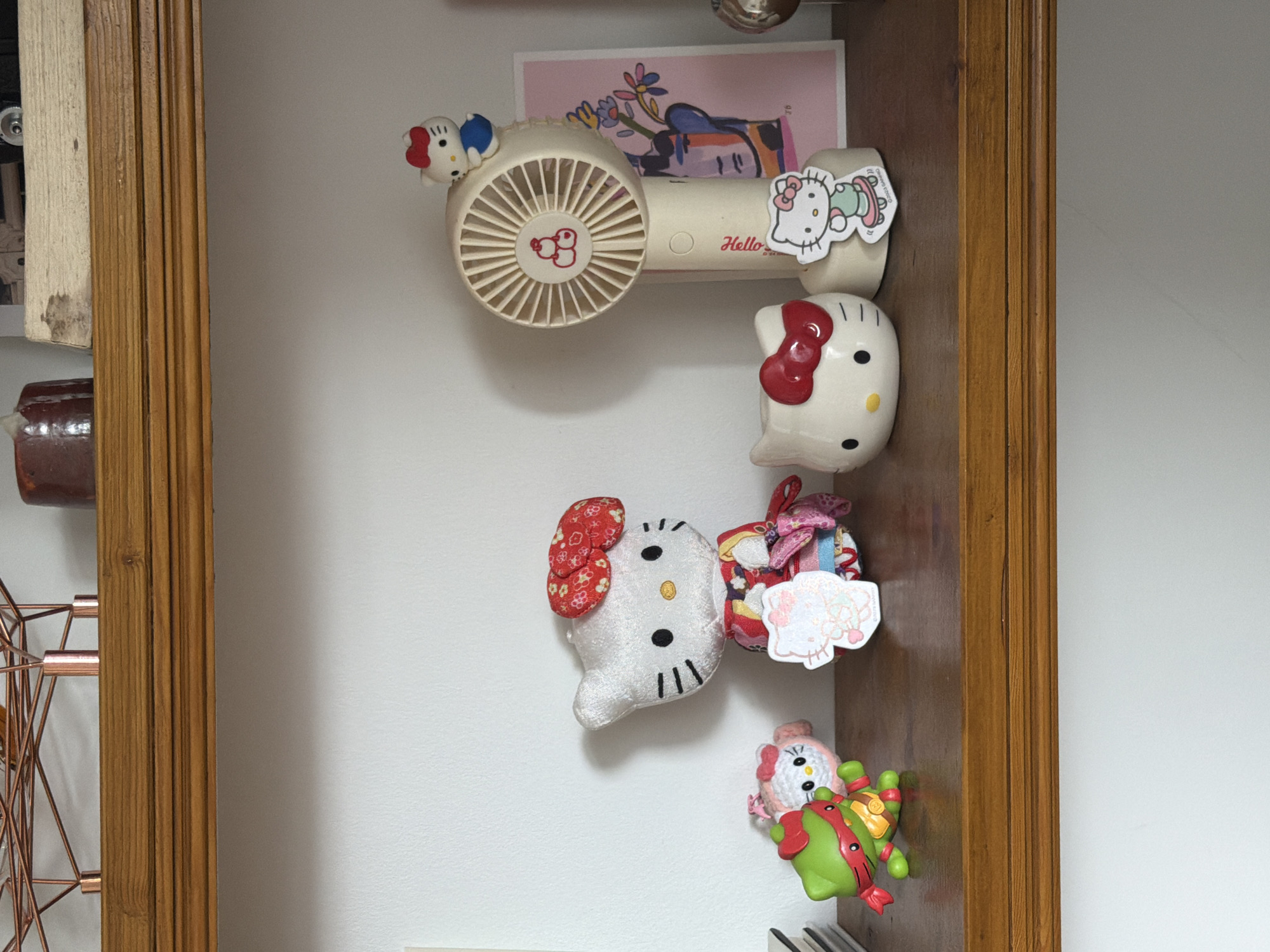 The image shows a collection of items within a wooden-framed space, possibly a shelf or cabinet, against a light-colored wall. The objects are arranged vertically, with several Hello Kitty-themed items dominating the display. These include plush toys and possibly decorations. Above them is a small electric fan with Hello Kitty stickers attached. A pink card is visible behind the fan. At the bottom is a Teenage Mutant Ninja Turtle toy. The presence of these items suggests a personal or domestic environment. The location of Folkestone and Hythe District, United Kingdom, is inferred by image metadata.