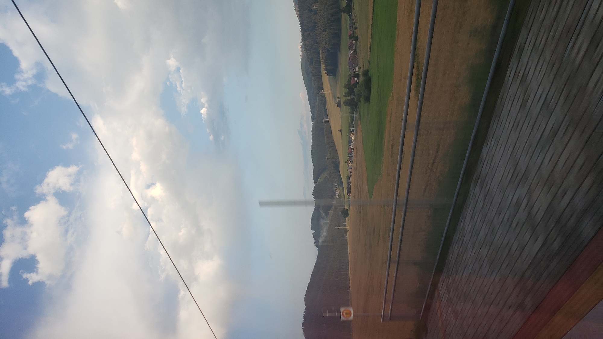 The image depicts a rural landscape in Lučivná, Slovakia, as seen from what appears to be a moving train. In the foreground, a wooden platform is visible, with a metal railing running along its edge. Beyond the railing, a vast field of golden-brown crops stretches towards a gently rolling hillside. The hillside is covered with a mix of trees and open, grassy areas, with some structures and power lines dotted across the landscape. In the middle ground, a small cluster of houses forms a village. The sky above is partly cloudy, with patches of blue sky peeking through. A thin wire stretches diagonally across the upper portion of the frame. The overall impression is one of serene natural beauty and agricultural activity in a Slovakian countryside setting.