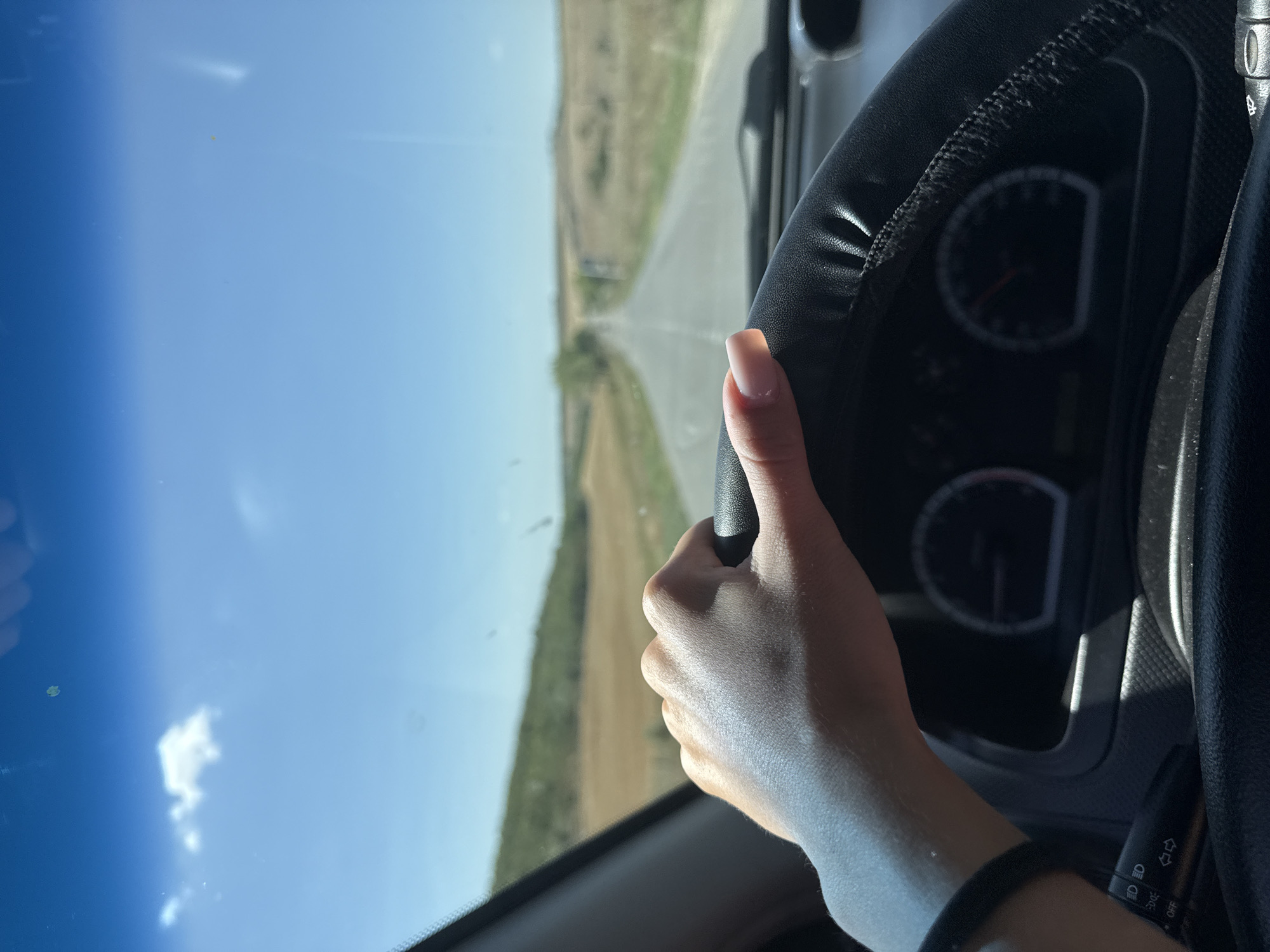 The image shows a first-person perspective of someone driving a car on a sunny day. The driver's left hand is on the steering wheel, which is covered in black leather. The steering wheel is positioned in the foreground, with a portion of the car's dashboard visible below it, including a speedometer. The view through the windshield reveals a long, empty road stretching into the distance, with dry, grassy fields on either side. Rolling hills, covered in sparse vegetation, are visible in the background. The sky is a clear blue with a few small white clouds. There is no indication of the city of Basarabeasca, Moldova, in the image.