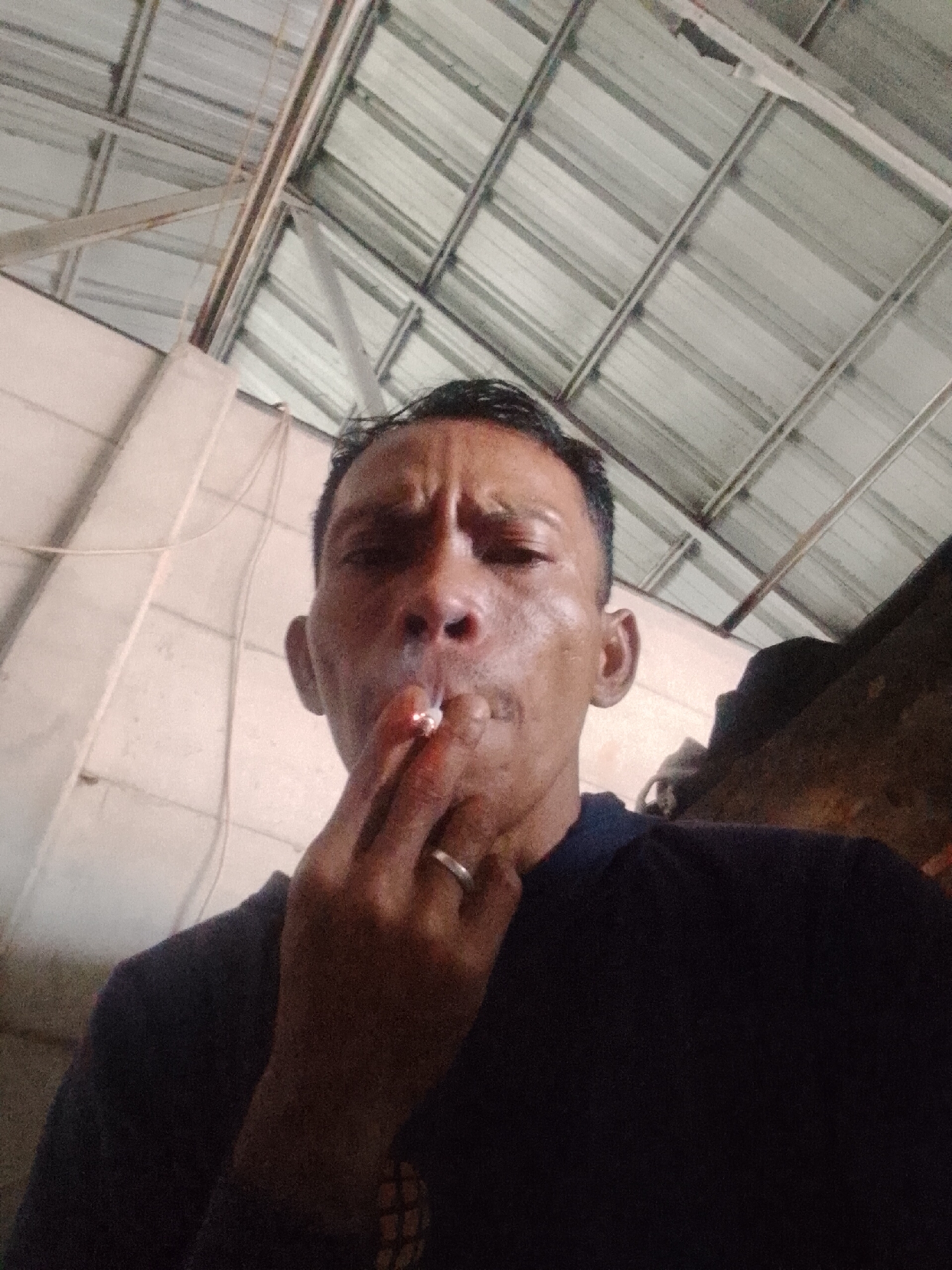 The image depicts a man smoking a cigarette. He has short, dark hair and is wearing a dark-colored shirt. A silver ring is visible on his finger. The background features a corrugated metal roof and white walls. The location is Bukit Ketok, Indonesia.