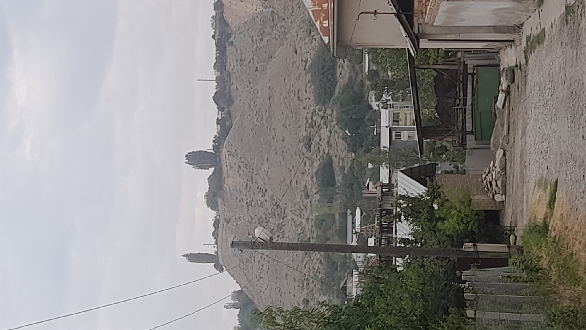 The image shows an outdoor scene with a dirt road leading towards a hill in the background. The hill is covered in dry vegetation and appears to be a significant geological feature. In the foreground, there are several buildings, including a house with a rusted metal roof on the right and a fence on the left. There are also piles of stones and construction materials on the side of the road. Trees and foliage are visible throughout the scene. The sky is overcast, suggesting a cloudy day. There are no people visible in the image. The location is described as Unknown, Unknown.