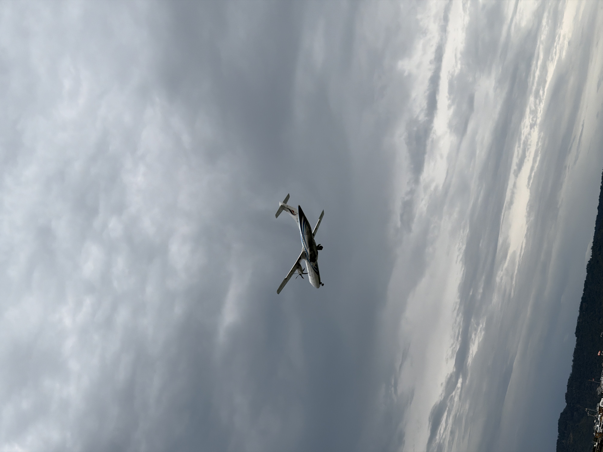 An airplane is seen flying against a cloudy sky in Ko Samui District, Thailand. The aircraft is a twin-engine turboprop, white with blue and red markings on its tail. It is mid-flight, with its propellers visible. The sky is overcast with varying shades of grey clouds. In the lower part of the image, a dark, hilly landscape is visible, possibly representing a coastline or mountains. There are some faint structures or lights visible on the very bottom edge of the image, suggesting it might be near a town or port. There are no people or distinct interactions visible.