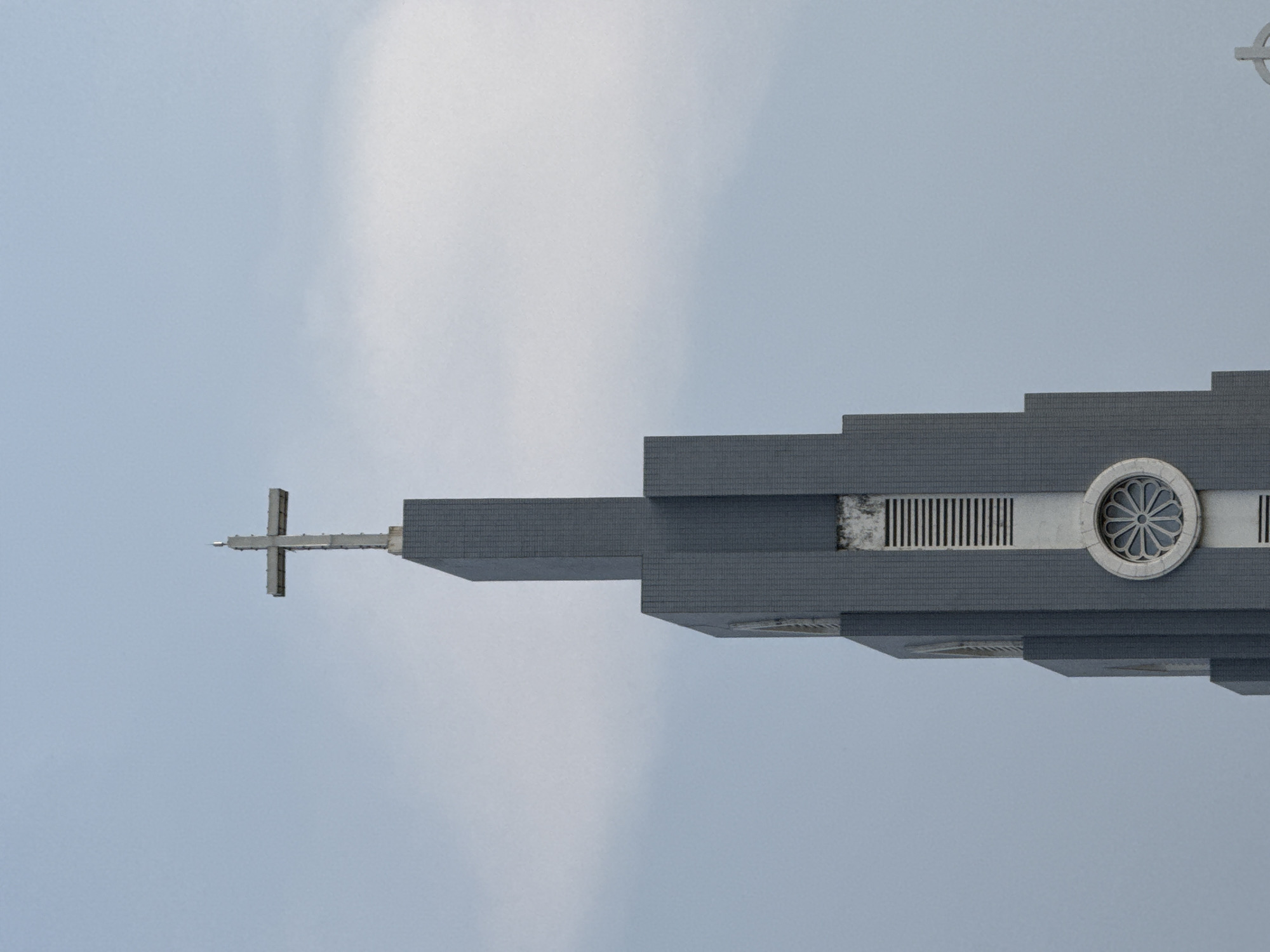 The image shows the steeple of the Thủ Đức Church in Thủ Đức, Vietnam, viewed from below. The steeple is gray and built in a stepped design with a cross at the top. Architectural features include a round window with a floral design and a series of vertical vents. The background is a cloudy sky.
