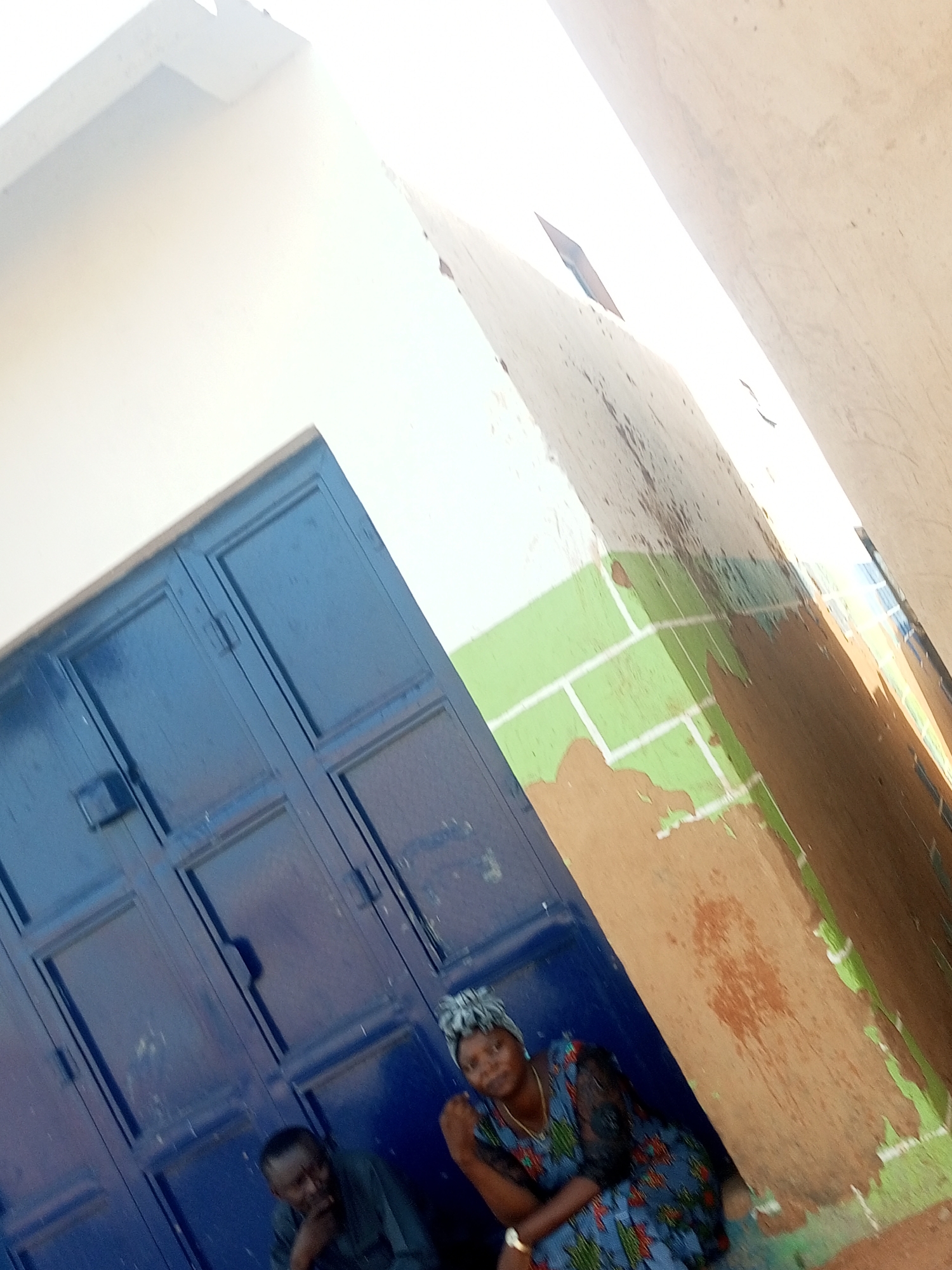 A photo taken at an extreme angle shows a man and woman sitting near a dark blue metal door and a wall painted green and brown in the city of Gombe, Nigeria. The wall has areas where the paint is peeling. The woman is wearing a dress with a floral pattern, while the man is in dark clothing. The top portion of the photo is washed out by sunlight.