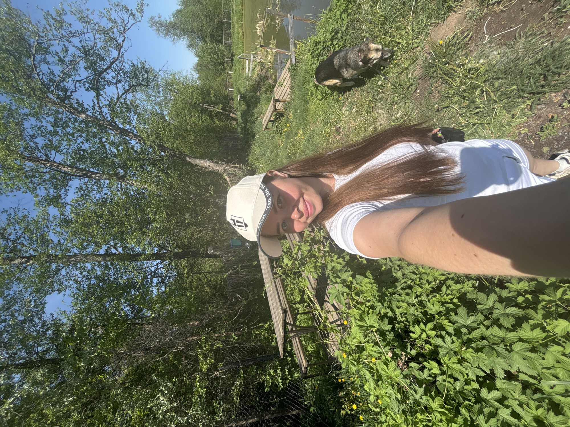 A woman wearing a white T-shirt and baseball cap is taking a selfie outdoors in Malyi Bukor, Russia. The background includes trees, a grassy area with yellow flowers, and a body of water with a small wooden structure. A dog is also present in the grass. A wooden bench is partially visible in the foreground.