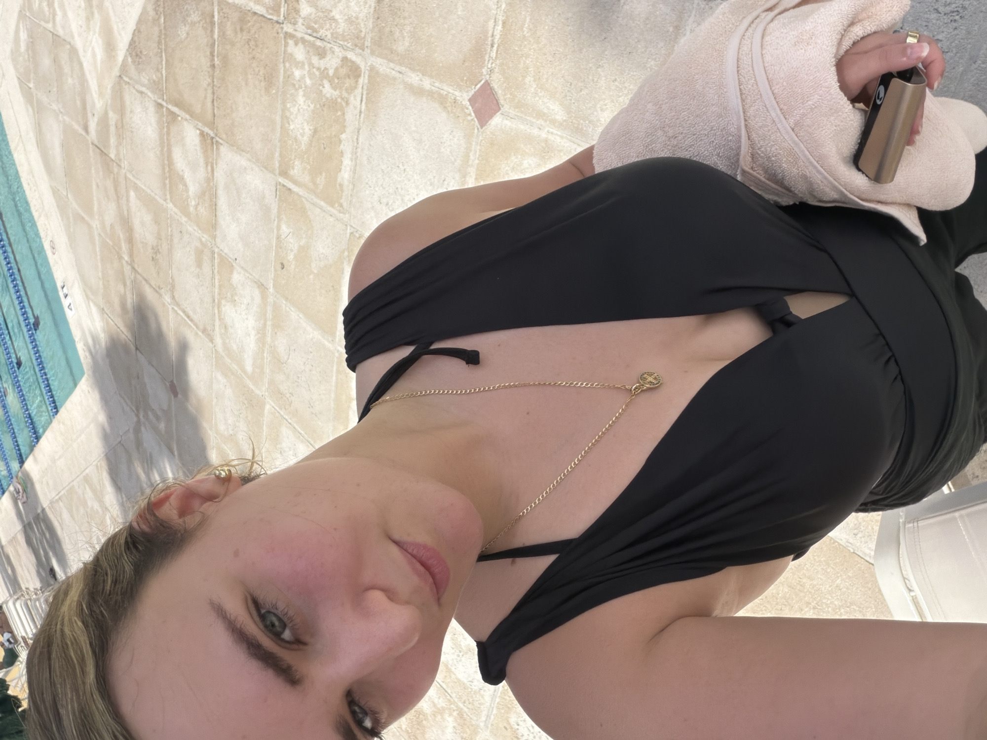 A medium, eye-level, rotated, and extreme close-up shot captures a young woman with fair skin and blonde hair, seemingly at a poolside in Key Biscayne, Florida. The woman is wearing a black bathing suit or a dress-like garment with a revealing neckline and a gold necklace. She is holding a beige towel in her right hand, which also holds a black and bronze-colored device. In the background, a tiled pool area is visible. There is a chair with a white back support slightly behind the woman. The angle of the shot is canted, giving the impression that the woman is holding the camera above her and to the side.