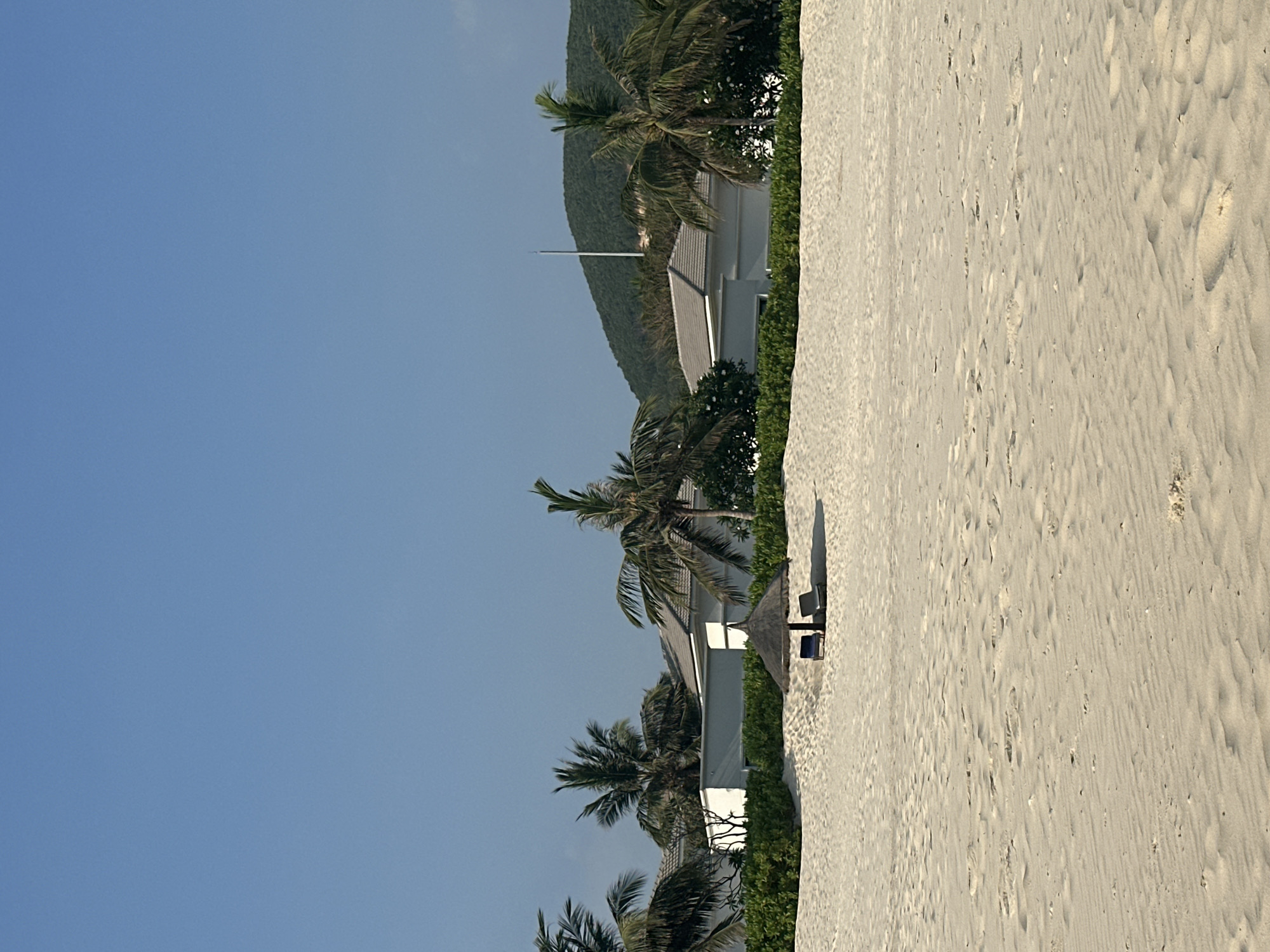 The image displays a serene tropical or coastal scene under a clear, bright blue sky. The perspective is along a wide expanse of light-colored sandy beach. To the left, a row of white resort-style buildings with grey roofs is visible, nestled behind lush green foliage and several tall palm trees. A prominent white flagpole stands erect behind one of the buildings.
In the foreground and mid-ground of the sandy beach, there is a straw-thatched beach umbrella positioned over two dark-colored lounge chairs. These objects cast distinct shadows on the sand, indicating bright sunlight and suggesting the time of day is around midday or early afternoon. The sand itself shows natural undulations and patterns, likely from wind or previous footsteps. No people are visible in the scene, contributing to a sense of tranquility and an inviting, uncrowded atmosphere. There is no visible text in the image.