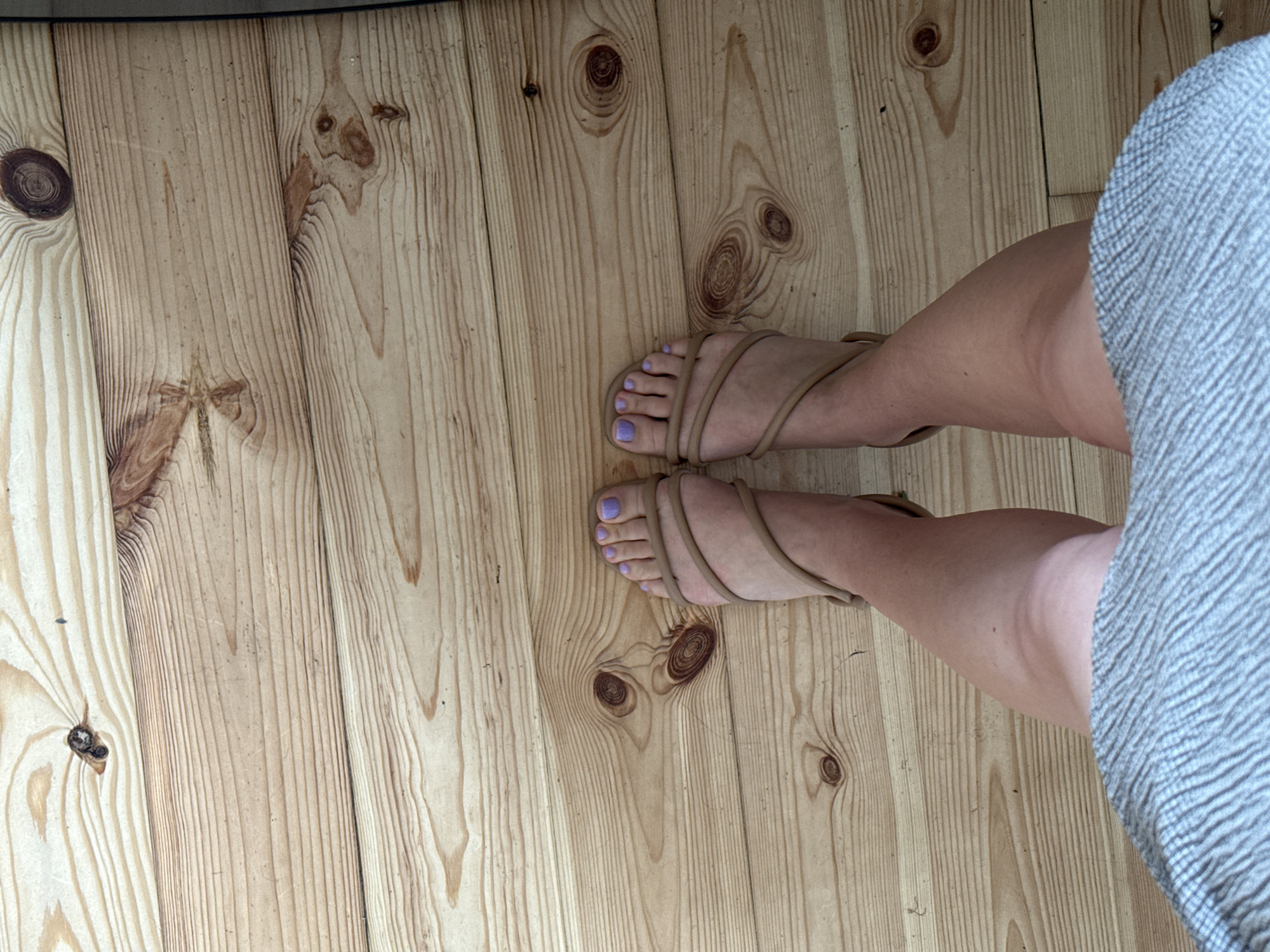 The image shows a person's legs and feet wearing tan strappy sandals with lavender-painted toenails, standing on a light-colored wooden floor. The floorboards are oriented horizontally and show wood grain and knots. The person is wearing a light gray and white patterned skirt that covers their lower legs and thighs. The scene is indoors, with no indication of a specific location in Łódź, Poland.