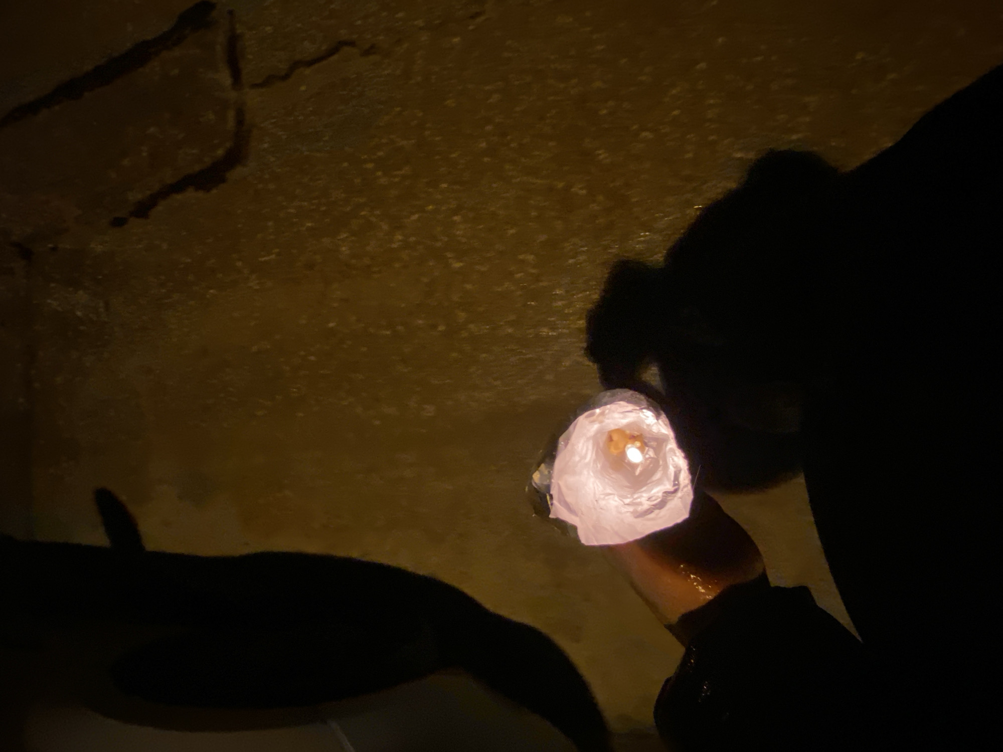 The image shows a hand holding a glowing, makeshift lantern inside what appears to be a dark, possibly underground, location. The light from the lantern illuminates part of the wall, revealing a rough, uneven surface. The person holding the lantern is mostly obscured by shadow. There is no identifiable information in the image that specifically locates it in Konstantinovo, Bulgaria.