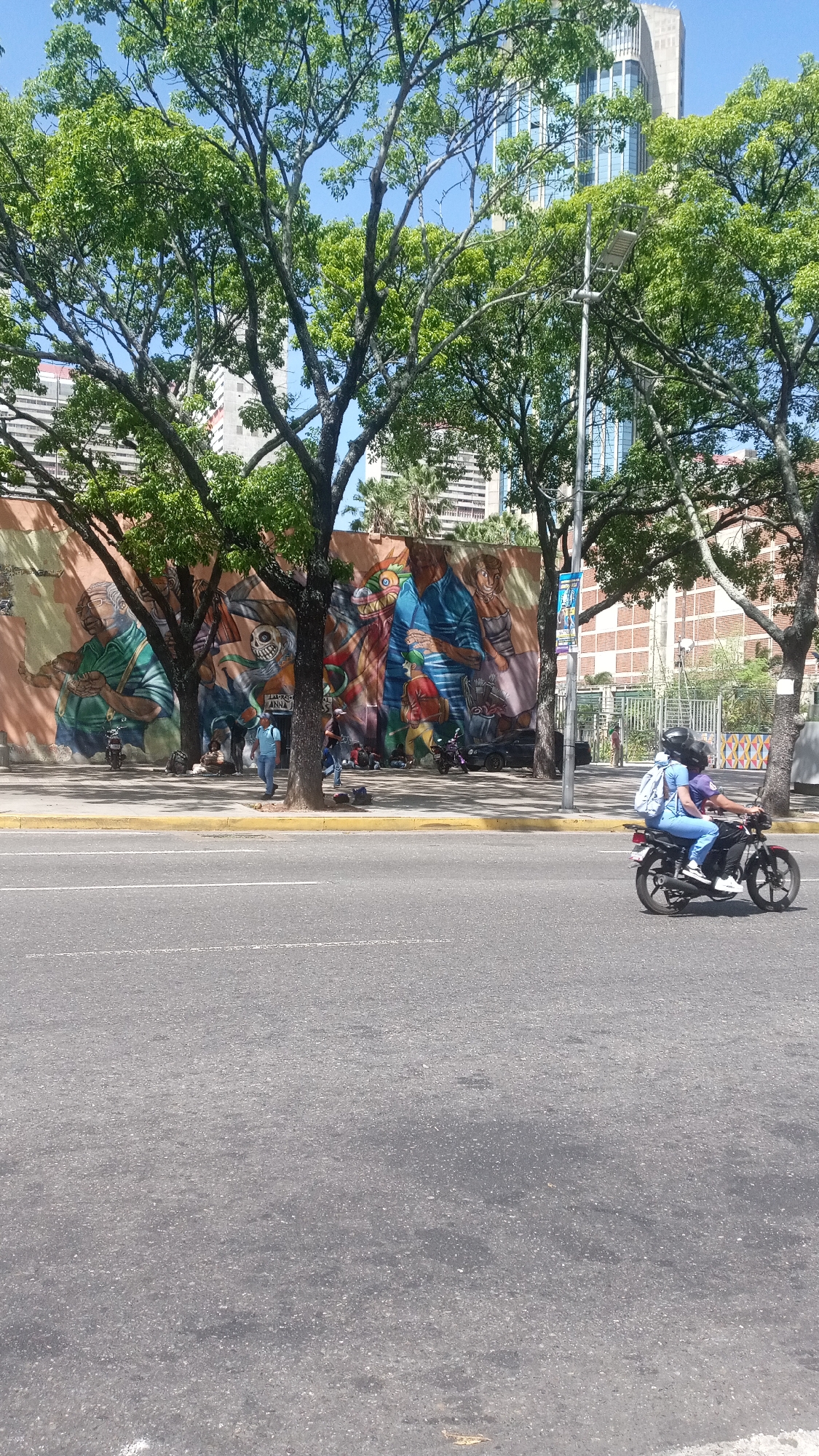 Here is a detailed description of the image:
**Location:** The scene appears to be an urban street in Unknown, Unknown. There is a long mural painted on a wall that runs alongside the sidewalk. Buildings, including a taller skyscraper, are visible in the background.
**People:** Several individuals are present in the image. Some are sitting or standing near the mural on the sidewalk. Two people are riding on a motorcycle that is moving along the street.
**Objects:**
- Mural: A large, colorful mural is a prominent feature of the scene.
- Trees: Several trees are planted along the sidewalk, partly obscuring the mural and surrounding buildings.
- Street: A wide street with a white line marking lanes is in the foreground.
- Motorcycle: A motorcycle with two people is moving along the street.
- Buildings: Buildings of different sizes and styles are in the background, including a skyscraper.
- Streetlight: A streetlight is visible along the sidewalk.
**Actions/Interactions:** People are walking, sitting, and interacting on the sidewalk. Two people are riding a motorcycle on the street.
**Environment:** The environment is urban, with trees, buildings, and a street. The lighting suggests it is daytime.