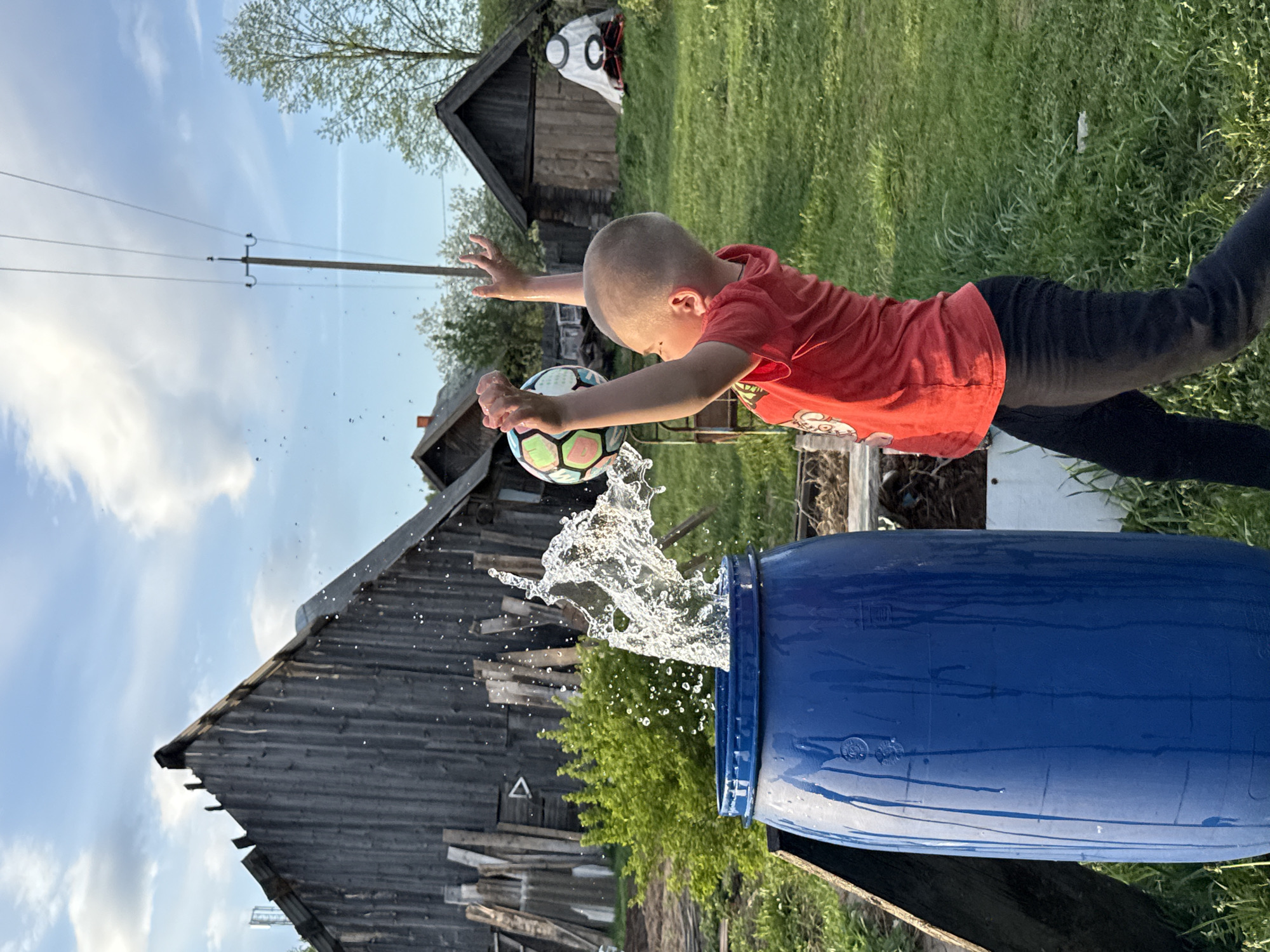 A boy in a red shirt and black pants is suspended mid-air, reaching out to a soccer ball positioned above a large blue plastic barrel. Water is splashing upwards from the barrel, suggesting the ball has just made contact with the water inside. In the background, there are dilapidated wooden buildings, a utility pole, and greenery. The sky is partly cloudy. The location is Unknown, Russia.