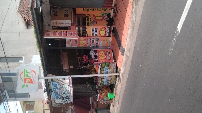 The image depicts a street scene in Kadipaten, Indonesia, located at coordinates -7.125 latitude and 108.146 longitude. It features a street lined with several small businesses, one of which is a fruit stand with a sign that reads "Dewi Fresh Fruit." There are several signs in Indonesian indicating the sale of "Bakso Oren," a type of Indonesian meatball soup. The fruit stand is decorated with a variety of fruits including oranges, lemons, and limes. The street is paved and there is a white line painted along the side of the street. There is a blue plastic bucket on the sidewalk in front of the fruit stand. The street is empty and there are no people in the scene.