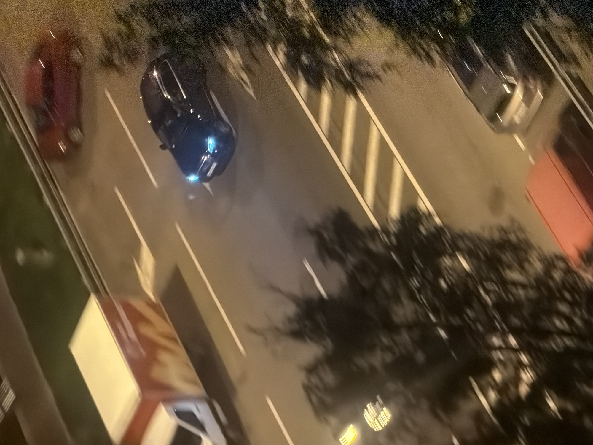 This is a high-angle, slightly blurry, nighttime aerial photograph of a street scene in Moscow, Russia. The image captures several vehicles in motion.
On the left side of the image, a white delivery truck with an orange and white insulated cargo box is driving on the road. The cargo box appears to have a graphic design on its side.
Further down the road, a dark-colored sedan, possibly black or dark blue, is moving from top to bottom in the frame. Its headlights are illuminated, creating a glow on the road. Behind it, a red sedan is also visible, also in motion.
In the lower part of the image, a silver SUV or crossover vehicle is parked or moving slowly on the right side of the road. To the left of this vehicle, a red object, possibly the roof of another vehicle or structure, is visible.
The road has clear white lane markings. There are trees with dark foliage casting shadows on the road and sidewalk area. Some yellow lights, possibly from streetlights or traffic signals, are visible in the background. The overall environment suggests an urban setting at night.