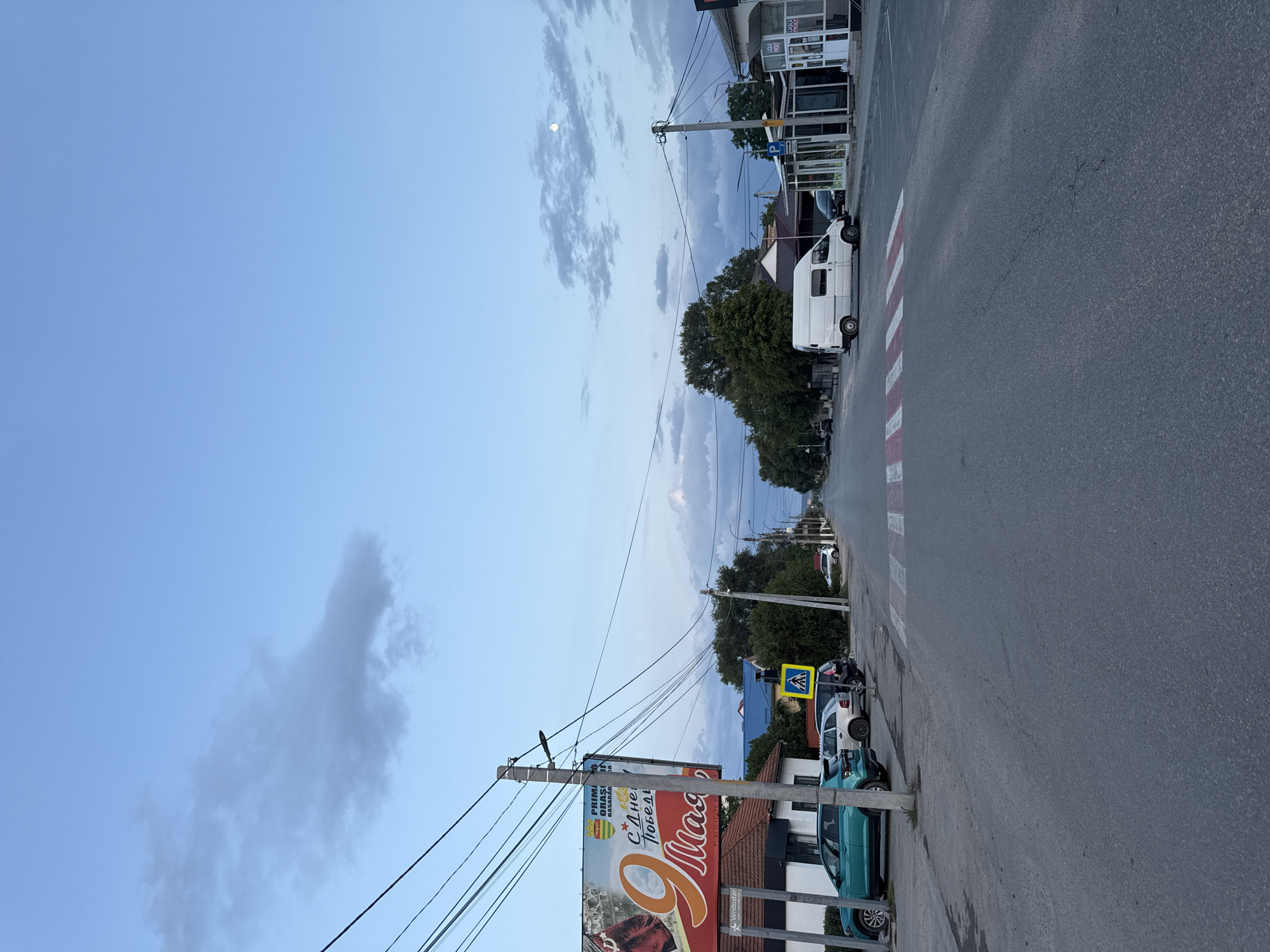 The image depicts a street scene in the city of Basarabeasca, Moldova, during dusk. The sky is a pale blue with scattered clouds and a visible full moon. A power line runs diagonally across the upper left quadrant. On the left side of the street, a large billboard with Cyrillic text and an image of the number "9" is prominently displayed above a building with a red-tiled roof. Several cars, including a teal hatchback and a silver SUV, are parked along the side of the road.
A pedestrian crossing with red and white stripes is marked on the asphalt in the center of the street. Further down the road, on the right side, is a white van parked parallel to the curb. Adjacent to the van are storefronts with glass windows and signage, including "LIQUI MOLY." Trees line both sides of the street, with more buildings visible in the distance, suggesting a moderately developed urban or suburban area. The overall impression is of a quiet street at the end of the day.
