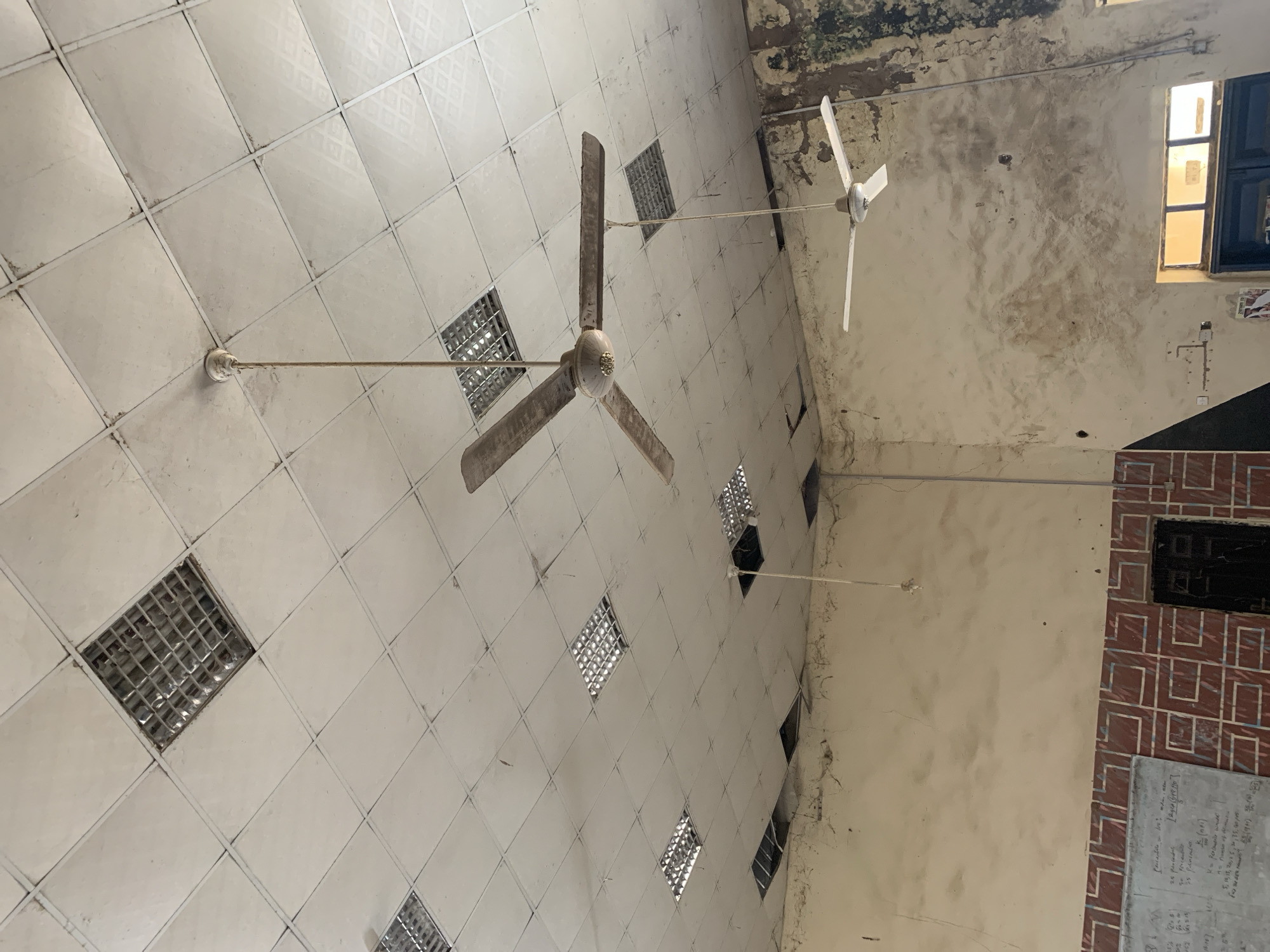 A wide-angle, eye-level shot captures the interior of a dimly lit room, possibly a classroom or lecture hall, in Jalingo, Nigeria. The ceiling, a grid of white tiles, shows signs of disrepair with several square light fixtures and some tiles missing, revealing darker openings. Two ceiling fans are visible: a larger, older model with wooden blades hangs centrally, suspended by a thick cord, while a smaller, modern white fan is mounted closer to the right wall. The walls are a muted, textured beige, stained and peeling in places, particularly on the right side, revealing patches of darker, possibly moldy, material. A portion of a whiteboard with handwritten equations is visible on the left. A dark-colored door is partially visible on the lower left. The overall impression is one of neglect and disuse.