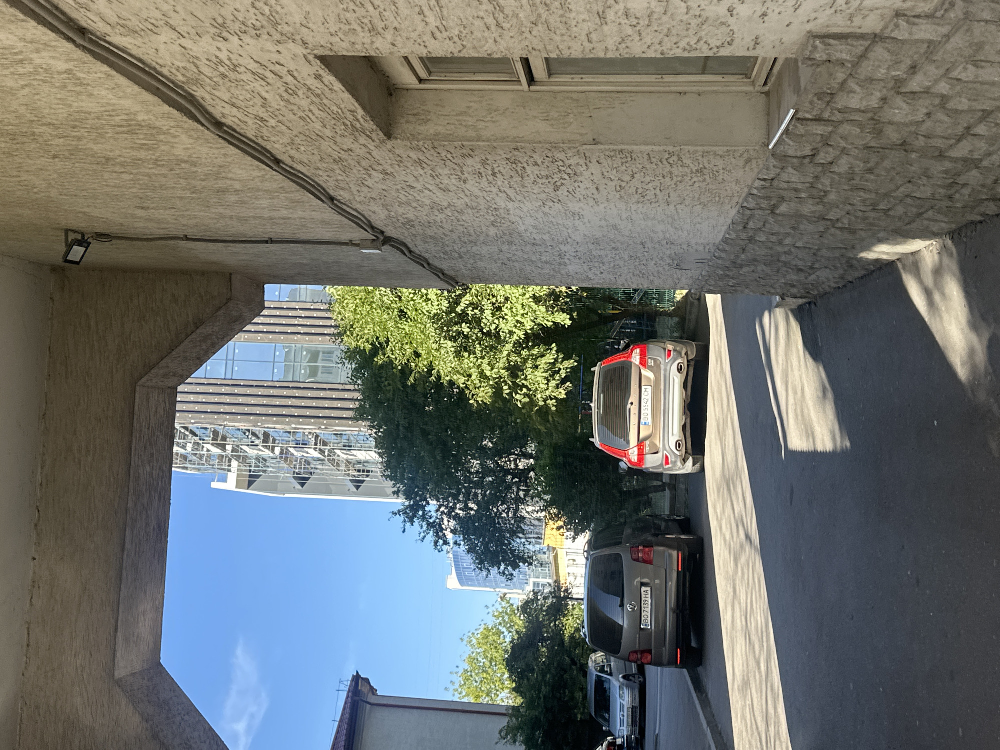 This is a ground-level, outdoor, daytime photograph taken in Ternopil, Ukraine. The perspective is from under an arched overhang, looking out towards a street scene bathed in sunlight.
In the foreground, on the right, is a textured, light-colored building with a stone foundation. A window is visible on this building, with a small metal object resting on the windowsill. Electrical conduits run along the building's facade, leading to a security light mounted on the overhang.
The street in front of the building has two cars parked, facing away from the camera. The car on the left appears to be a grey Volkswagen Touran, with a license plate reading "BO 7139 HA". To its right is a Volvo XC60, also grey, with a license plate reading "BO 5542 CM". A third silver car is partially visible further down the street on the left.
Behind the parked cars, there are green trees and a tall, modern building with a glass facade under construction, surrounded by scaffolding. The sky is clear and blue. The ground in front of the cars is asphalt, with distinct shadows cast by the cars and trees.