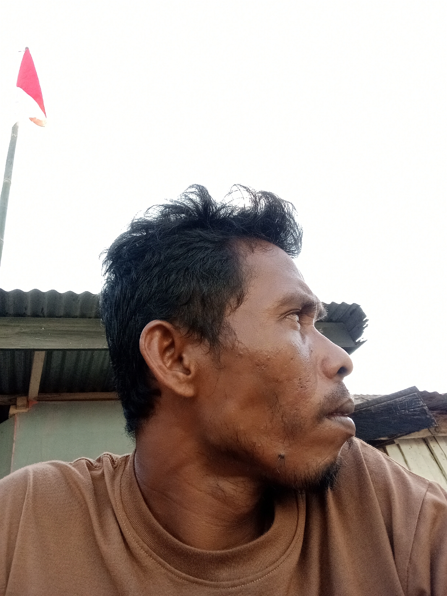 A man's head and shoulders are captured in a close-up, profile shot, looking to the right. He has dark, tousled hair and is wearing a plain brown t-shirt. In the background, a red and white flag flies on a pole to the left. Further behind, corrugated metal roofing is visible, suggesting a simple, possibly rural, structure. The sky is bright and overcast. The image was taken in Lakea Satu, Indonesia.