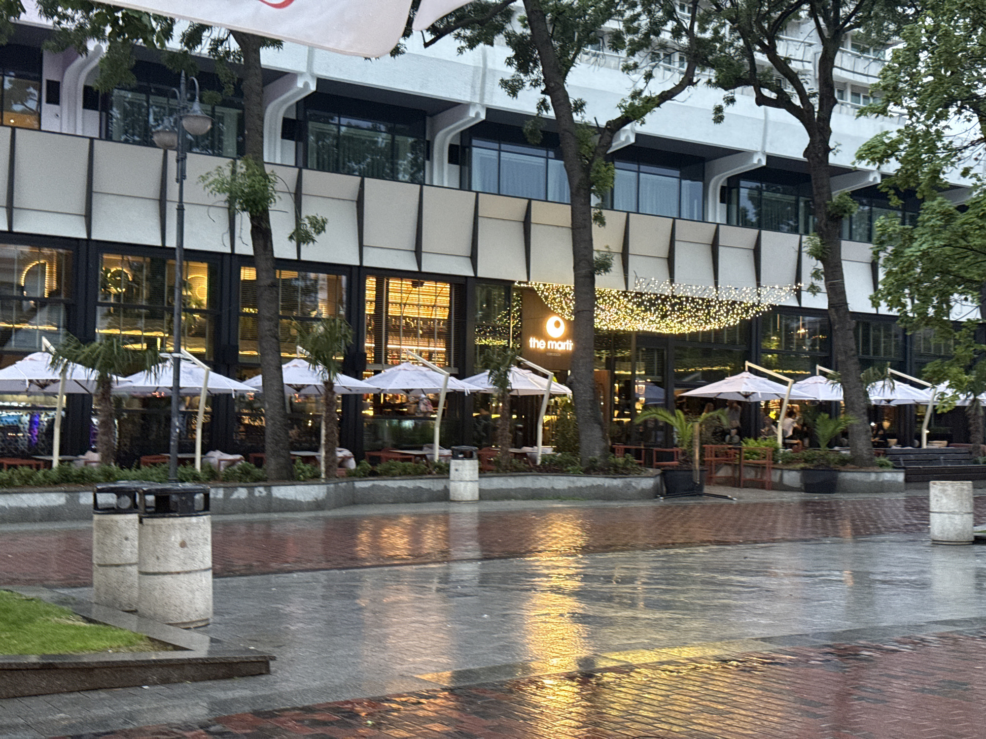 This image depicts an urban street scene in Varna, Bulgaria, captured likely in the late afternoon or early evening after a recent rain shower. The foreground consists of a wet, reflective paved area, shimmering with light reflections. The pavement includes sections of brick-like patterns and smoother grey surfaces. On the left, two cylindrical concrete public bins with black tops are visible near a patch of green grass.
In the midground and background, a modern, multi-story building houses ground-floor commercial establishments with expansive glass windows, revealing warmly lit interiors. An outdoor dining area extends along the sidewalk in front of these businesses, featuring numerous white umbrellas, tables, and chairs, with a few people faintly visible within. Palm trees and other plants in planters contribute to the ambiance. A prominent sign on one of the establishments clearly displays the text "the mart" next to a circular logo. Above this entrance, a canopy of decorative string lights casts a festive glow.
The upper floors of the building exhibit contemporary architecture with large windows and a combination of white and grey facade elements. Several tall, mature trees with dark trunks and green foliage are spaced along the sidewalk, partially obscuring the building and the lamppost in the foreground. The wet ground reflects the artificial lights, creating a serene yet inviting atmosphere, suggesting a quiet moment before bustling evening activity. A partial banner with a red logo is visible at the top of the frame.
