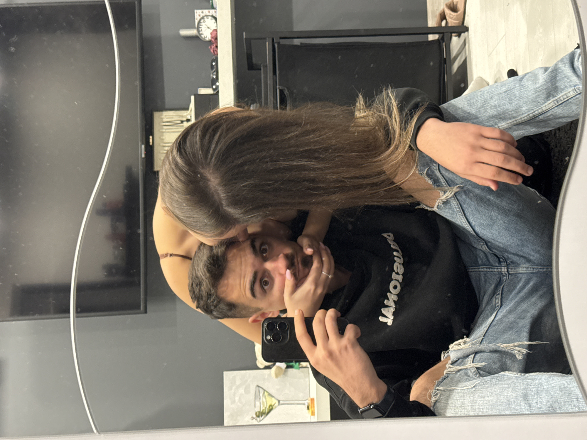 The image is a mirror selfie, presented on its side, showing a couple in an indoor setting. The man takes the photo, holding a black smartphone with a triple camera in his right hand. He is positioned with his torso angled upwards, wearing a black long-sleeved top with mirrored white text "JAWOIЯOWS" across the chest, and light blue ripped jeans. A black smartwatch is on his left wrist. His dark hair and beard are visible, and he looks directly at the camera with wide, surprised eyes. His left hand is raised towards his face, partly covered by the woman's hands.
The woman is leaning over him from above, her long, dark brown hair flowing downwards. Her face is close to his, and her hands gently cup his face. She is wearing a top with thin brown straps, partially visible.
The background, reflected in the mirror, includes a dark gray wall. On the left side of the frame, a large, dark flat-screen television is mounted. To the right, a wall-mounted clock with Roman numerals and some decorative items are visible. Below the TV, a picture of a martini glass hangs on the wall. The floor, towards the top of the frame, is light-colored, possibly wood or laminate, with a dark rug underneath the man. A black chair or furniture piece is also reflected. The scene conveys a playful and affectionate interaction between the couple.