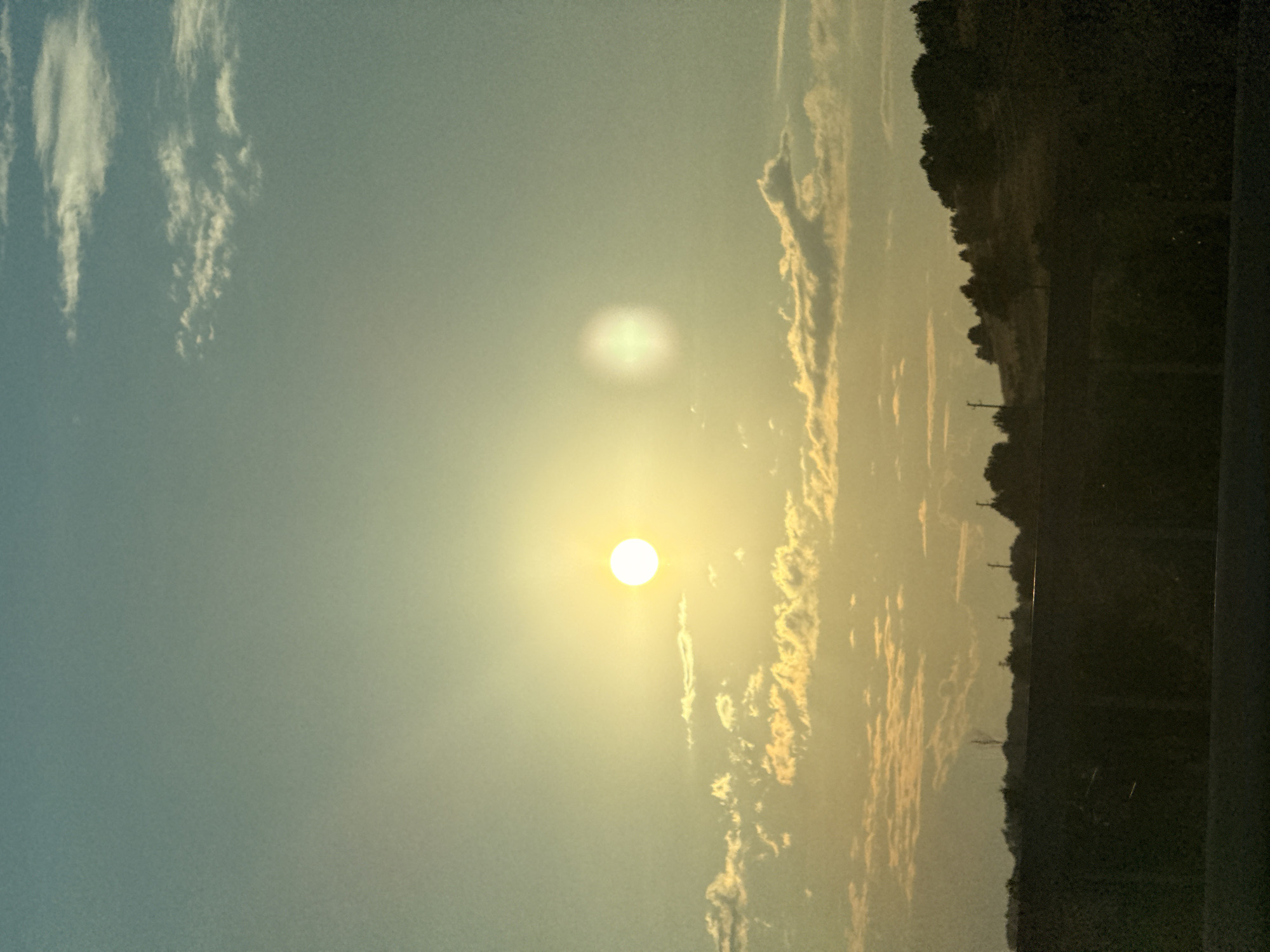 The image captures a serene sunset over Kilifarevo, Bulgaria. The sky is a canvas of soft blues and yellows, with wispy white clouds scattered across the expanse. The sun, a brilliant orb of light, is positioned slightly off-center, casting a warm glow that bathes the scene in a golden hue. In the foreground, a dark silhouette of trees and hills forms a dramatic contrast against the vibrant sky, suggesting a landscape setting. The overall mood is peaceful and tranquil, evoking the beauty of nature at dusk. No people or text are visible in the image.
