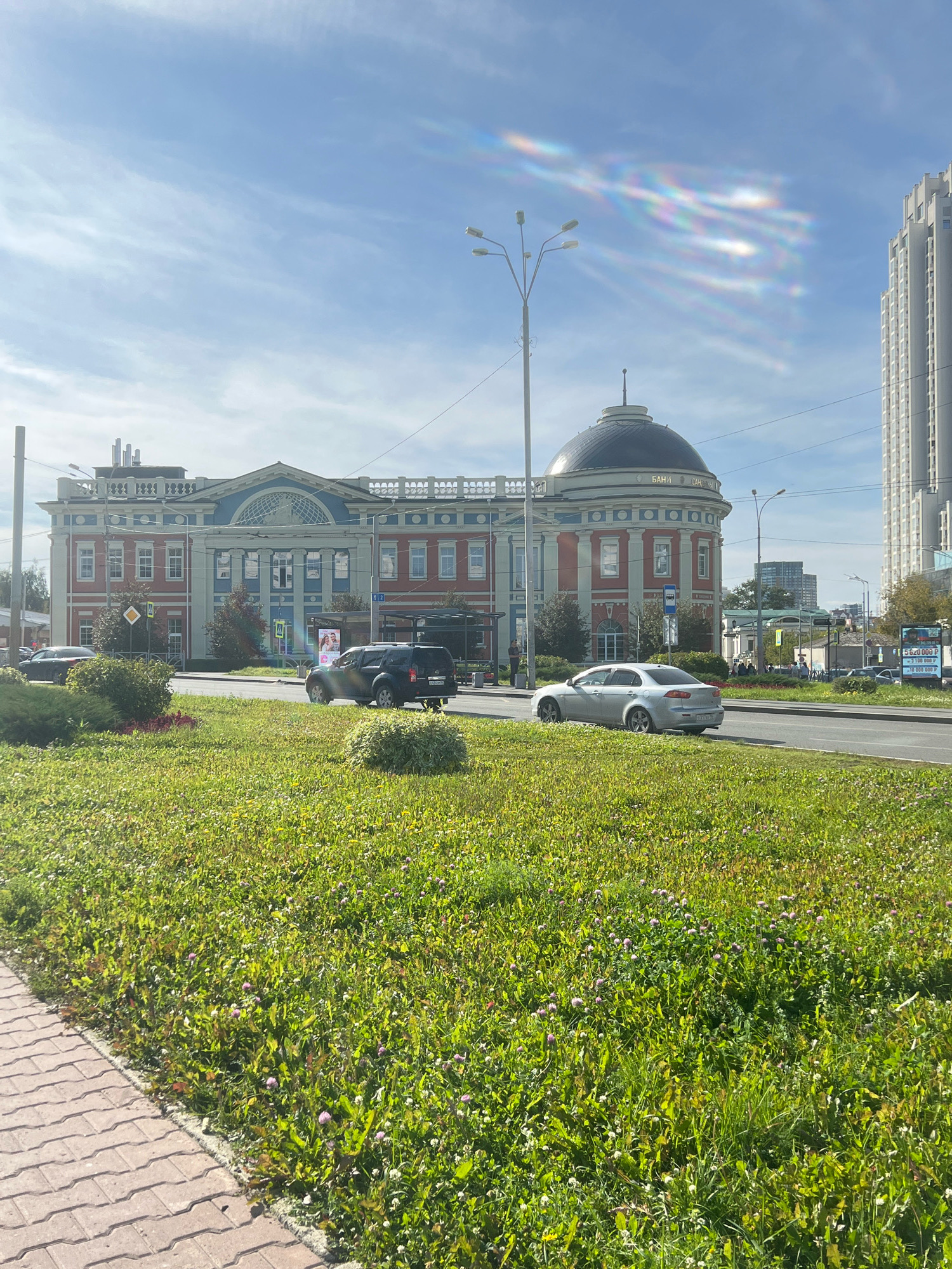The image shows a street scene in Yekaterinburg, Russia, with a large, ornate building with a dome as the central focus. The building, identified as the "Bank Saint-Petersburg" based on Cyrillic lettering visible on its facade, features classical architectural elements like columns and arched windows, painted in red and blue. To the right of this building, a modern, tall skyscraper rises, contrasting with the older architecture.
In the foreground, a wide median strip is covered in lush green grass with scattered pink and white wildflowers. A paved walkway with interlocking concrete blocks runs along the left edge of the image. A multi-lane road runs in front of the buildings, with a silver sedan and a dark SUV driving on it. Traffic signs and a bus stop are visible on the side of the road.
The sky above is blue with wispy white clouds, and there are visible rainbow-like light refractions, possibly from lens flare or atmospheric conditions. Streetlights are present along the road. The overall impression is a sunny day in an urban environment, showcasing a blend of historical and contemporary architecture.