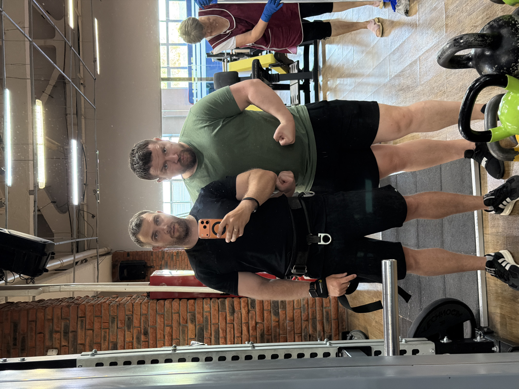 The image shows a mirrored selfie of two men in a gym.
The first man, wearing a black t-shirt and black shorts, holds an orange smartphone and takes the picture. He has short brown hair and a beard. He is wearing a weightlifting belt with a D-ring.
The second man, standing behind the first, wears a green t-shirt and black shorts. He is flexing his biceps.
In the background, an elderly woman is visible. She wears a burgundy top, black pants, and blue gloves, and appears to be cleaning gym equipment.
The gym contains various pieces of equipment, including weight machines, kettlebells, and a squat rack. The wall behind the selfie-taker features a brick design. The lights hanging from the ceiling are long and tube-shaped.
The location is identified as the city of Chaikovsky, Russia.
