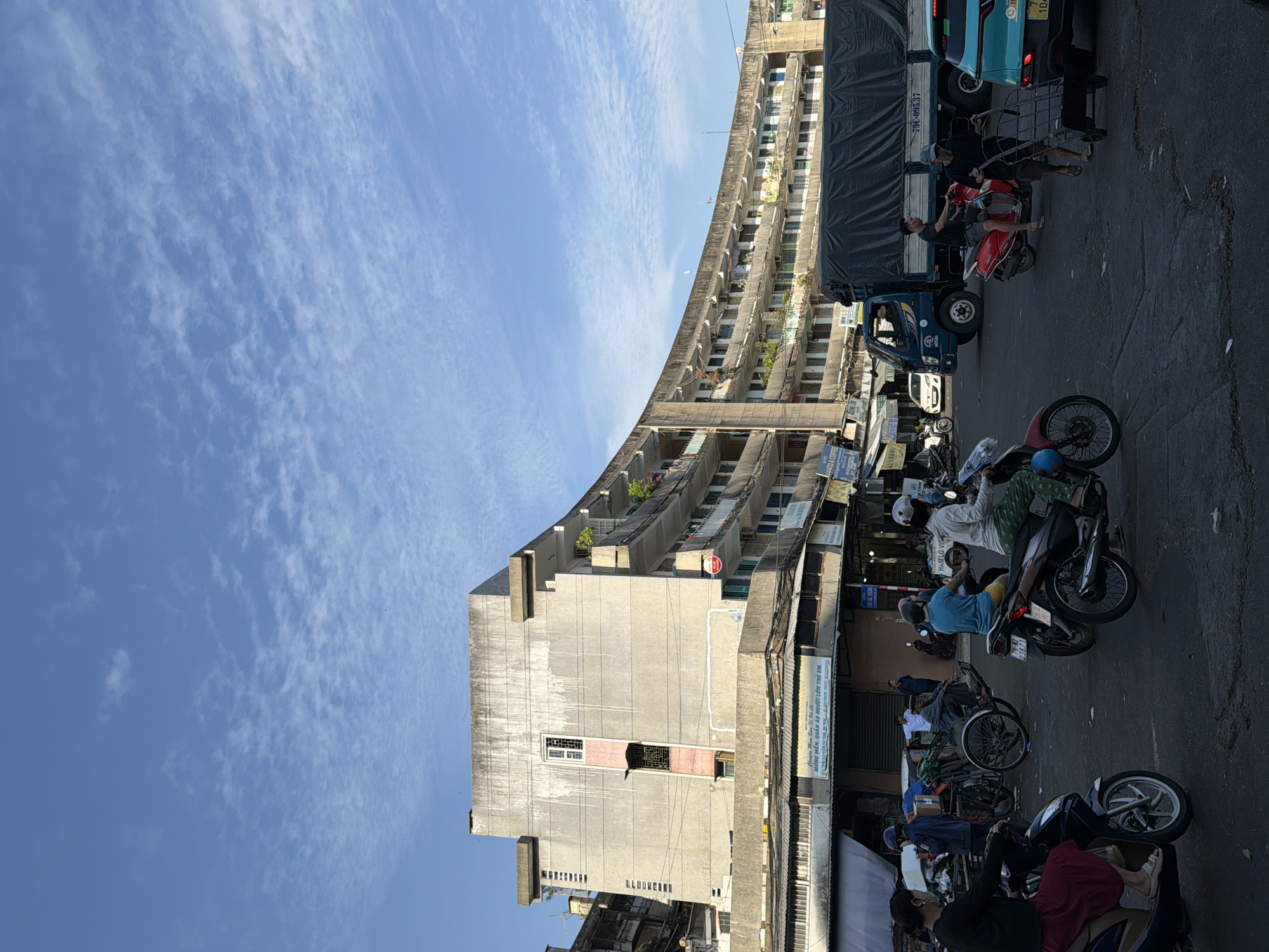 The image depicts a street scene in Nha Trang, Vietnam, under a blue sky with scattered clouds. In the background, a distinctive curved, multi-story concrete building with balconies dominates the view. The building shows signs of age, with visible stains and wear on its facade. Below the building, there are shops and stalls lining the street.
In the foreground and middle ground, several people are engaged in various activities. Many are riding motorcycles or scooters, some carrying passengers or goods. A large blue truck covered with a black tarp is parked on the right side of the street, its license plate partially visible as "79C-095.37". A teal-colored car is parked behind the truck, with a visible license plate containing "79A". A person is seen pushing a trolley loaded with items. Other individuals are standing or walking near the shops. The street surface appears to be asphalt and shows some signs of wear and tear. The overall atmosphere suggests a busy urban environment.