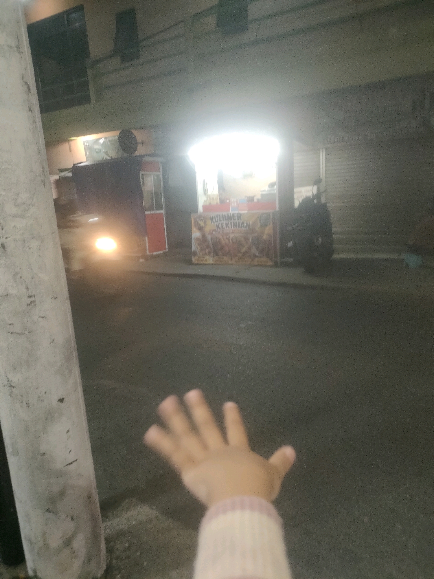 In a dimly lit street scene in Surabaya, Indonesia, a hand with spread fingers is visible in the foreground, partially obscuring a concrete pole. Behind the pole, a bright headlight from a moving vehicle illuminates the asphalt road. In the background, a food stall with the text "KULINER KEKINIAN" is visible, along with a motorcycle parked nearby. The surrounding buildings appear to be commercial establishments with closed shutters. The overall atmosphere is nocturnal and urban, suggesting a street-side dining or retail area at night.