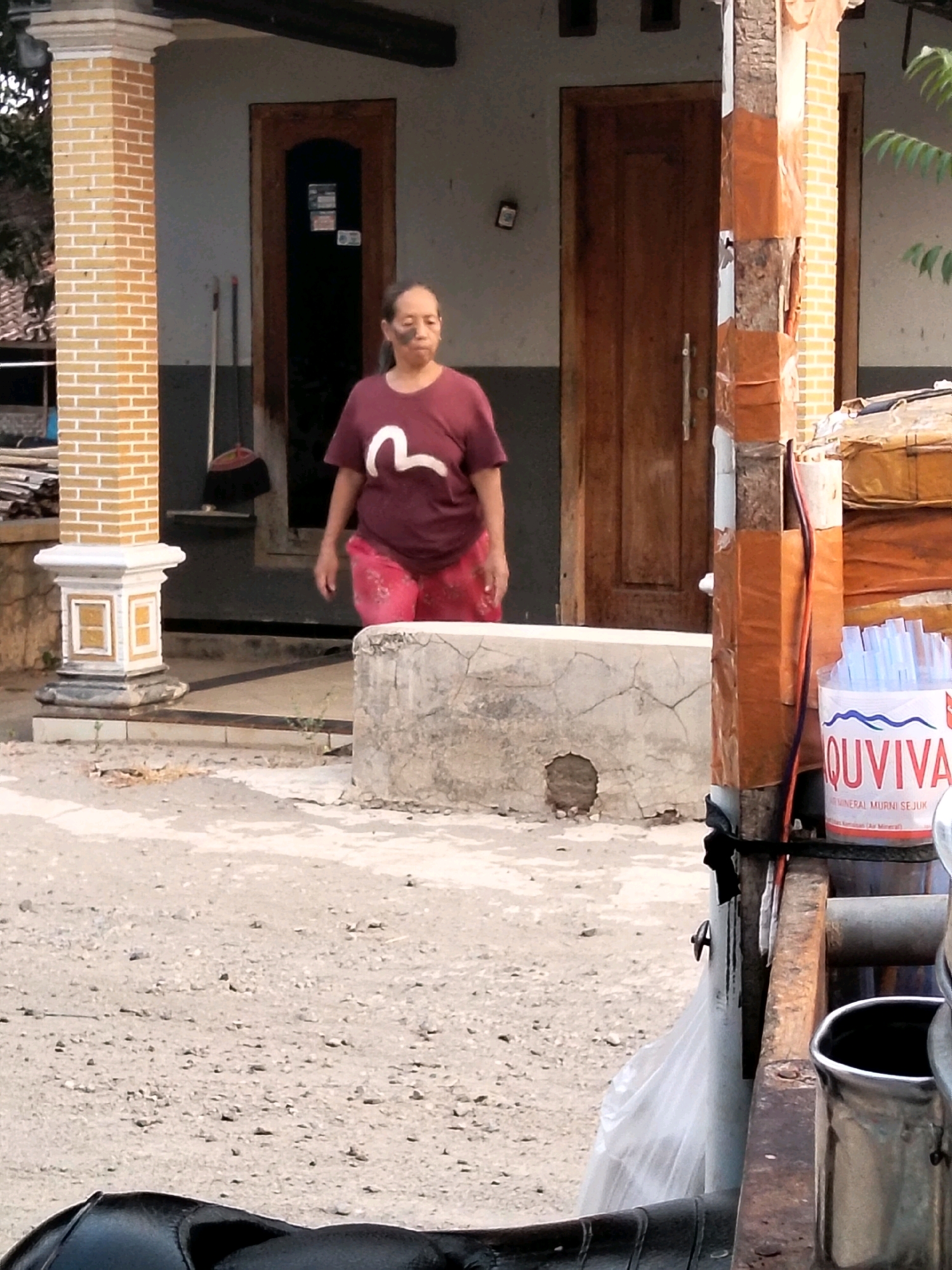 In the city of Jenangan, Indonesia, a middle-aged woman with dark hair is walking. She is wearing a maroon t-shirt with a white graphic and red patterned pants. The woman has a dark mark or stain on her cheek. She is standing on a dirt path in front of a building with a brick pillar and a wooden door. To the right of the woman, a cylindrical container holds numerous blue plastic straws. The container is labeled "QUVIVA". Behind the woman, a broom and dustpan are leaning against the building. The environment appears to be outdoor and slightly dusty.
