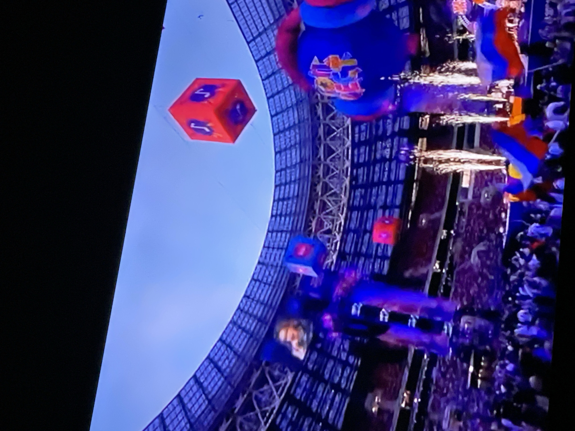 The image depicts a concert or sporting event taking place inside a large stadium, likely the Luzhniki Stadium in Moscow, Russia, due to the presence of Russian flags and the architectural style of the stadium's roof structure. A crowd of spectators is visible in the foreground, illuminated by stage lighting and pyrotechnics. In the background, there are large screens displaying images of performers, and several large, inflatable objects are suspended in the air, including a red cube with musical notes on its sides and a large blue object with colorful graphics. The event appears to be a lively and celebratory occasion.