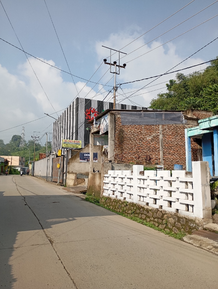 The image portrays an outdoor daytime scene on a paved road in the city of Situwangi, Indonesia, under a partly cloudy blue sky. The road, appearing to be asphalt or concrete with a visible central seam, extends into the distance, with shadows cast across its surface.
On the left side of the road, a large building with a distinctive black and white striped facade is visible, featuring a prominent red circular logo and several commercial signs, including yellow and white ones, near its base. Adjacent to this, an older structure with exposed red bricks and a corrugated metal roof is present, with posters affixed to its front. Further down the road on this side, a long, light-colored wall or fence, possibly part of a commercial property, is seen, accompanied by yellow utility boxes. A distant power transmission tower is visible on a hill beyond these structures. A white motorcycle is parked on the far left, and a dark-colored scooter with a single rider is observed further down the road, facing away.
On the right side of the road, a decorative white concrete block wall with an interlocking pattern rests upon a stone foundation, with green foliage growing at its base. Behind this wall, a brick building is partially visible, followed by another structure featuring a distinct blue painted section. Beyond these buildings, a dense area of green trees and vegetation covers a rising hillside. Overhead, multiple utility poles, including a prominent central one with a three-insulator cross-arm structure, support a complex network of electrical and communication wires that crisscross the sky.