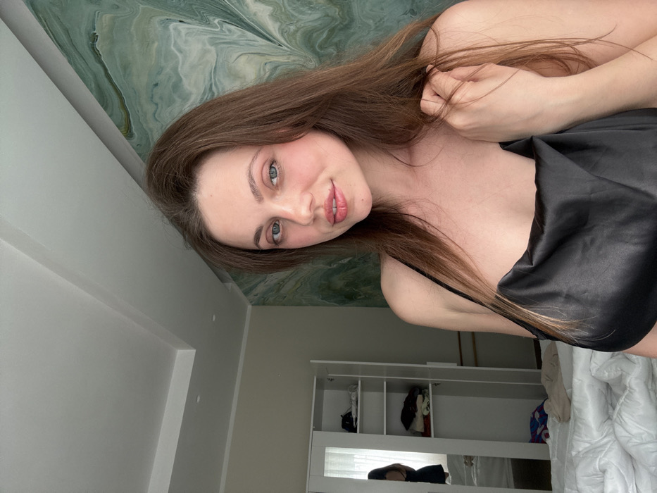 The image displays an indoor scene, likely a bedroom or dressing room. A female subject, appearing young to middle-aged with long brown hair and blue eyes, occupies the right half of the frame, facing forward and looking directly at the viewer with a slight smile. She is dressed in a black satin or silk camisole with thin straps. Her left hand is visible, gently touching her right shoulder area.
To the left of the subject, a white, two-door wardrobe is present. The left door is closed and reflective, showing a partial, indistinct reflection of a person from behind, wearing a dark garment. The right wardrobe door is open, revealing multiple interior shelves containing folded clothing items, with a black baseball cap visible on the top shelf. Below the wardrobe, a rumpled white duvet or comforter is partially visible, indicating a bed. The wall behind the wardrobe is plain light beige. To the right and behind the subject, the wall is adorned with a light green and white marble-patterned finish. The ceiling is white with a multi-level architectural feature. The lighting suggests natural light entering from an unseen source to the left. The image provides no discernible features or contextual information to confirm the location as Scafati, Italy.