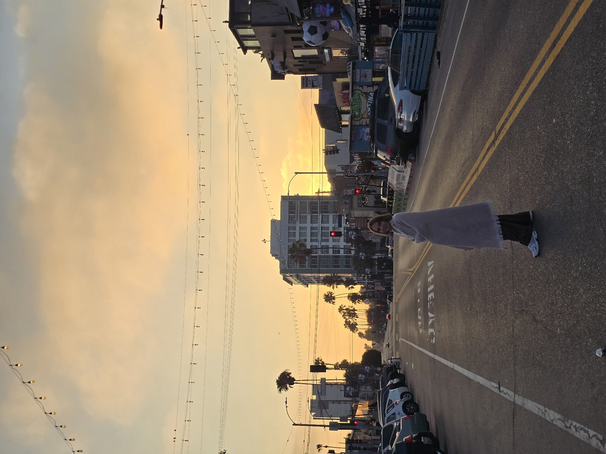 The image shows a street scene in Los Angeles, USA, during sunset. The sky is a vibrant orange and yellow, with strings of lights stretched across it. A woman is standing in the middle of the road, wrapped in a blanket. She is looking towards the camera. In the background, there are buildings, palm trees, and cars parked along the street. The street is paved and has yellow double lines in the center. The overall mood of the image is peaceful and serene.