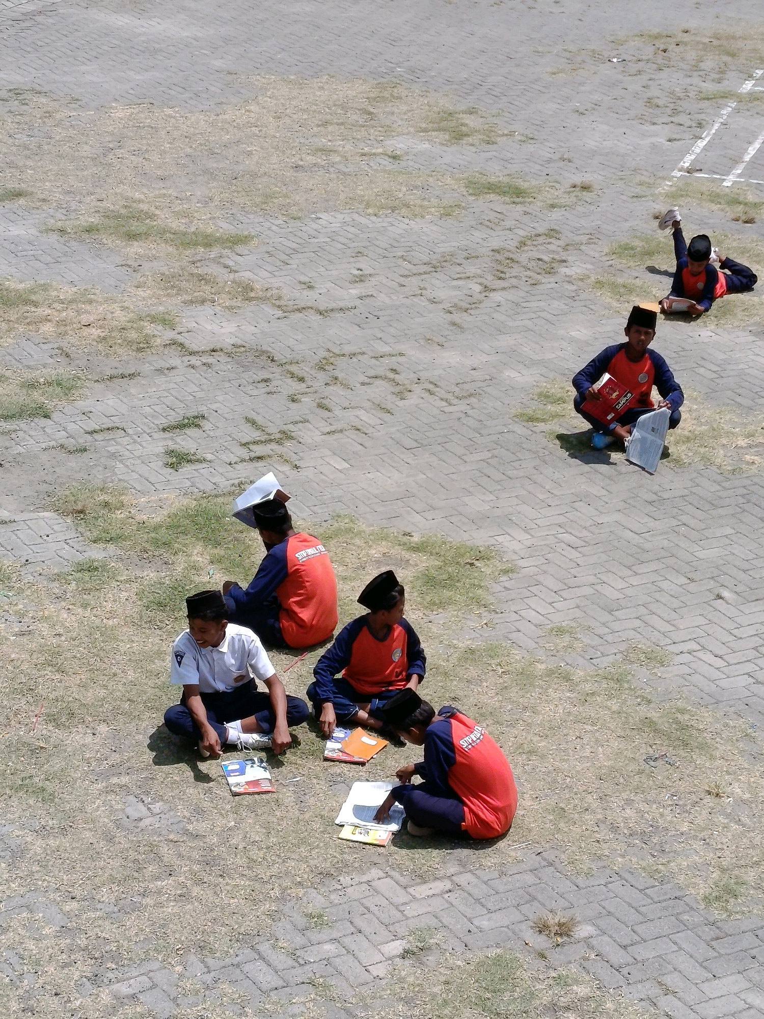 The image depicts an outdoor scene with several young people, presumably students, engaged in various activities on a paved and grassy area. In the foreground, two individuals are seated on the ground, facing the camera. One, wearing a white shirt and dark pants with a black cap, is looking to their right and appears to be smiling. In front of them are several books and papers. To their right, another individual, wearing a dark blue and red uniform with a black cap, is seated and looking down at a book, with more books and papers spread around them. Further back and slightly to the right, another student in a similar uniform is seated and reading a book. To the far left of the foreground group, a student in a blue and red uniform with a black cap is seated, holding a book over their head.
In the background, a student in a blue and red uniform with a black cap is lying on their back, reading a book, with their legs extended. Further in the distance, another student in a similar uniform is seen lying down, possibly also reading or resting. The environment consists of a gray paved area with rectangular blocks, interspersed with patches of dry grass and some scattered green tufts. White lines, potentially marking a sports field, are visible in the upper right portion of the image. The lighting suggests a bright, sunny day. The overall impression is of a school setting where students are taking a break or engaging in individual study outdoors.
