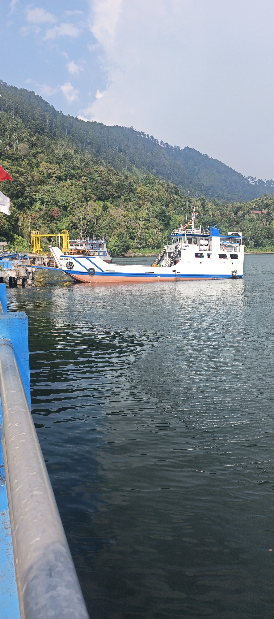 This is a medium-shot, eye-level, outdoor, daytime, full-color photograph taken at the pier in Nagaritongah, Indonesia. The image shows a ferryboat named "KMP SUMUT II" docked at a pier. The ferryboat is white with an orange stripe along its hull and a blue superstructure. The pier is also visible, with blue railings and metal supports. In the background, a lush green hillside rises steeply from the water's edge, covered with trees. The water is calm with gentle ripples reflecting the sky and the surrounding landscape. The sky is partly cloudy with patches of blue. The scene conveys a sense of tranquility and a typical day at a port in a tropical region.