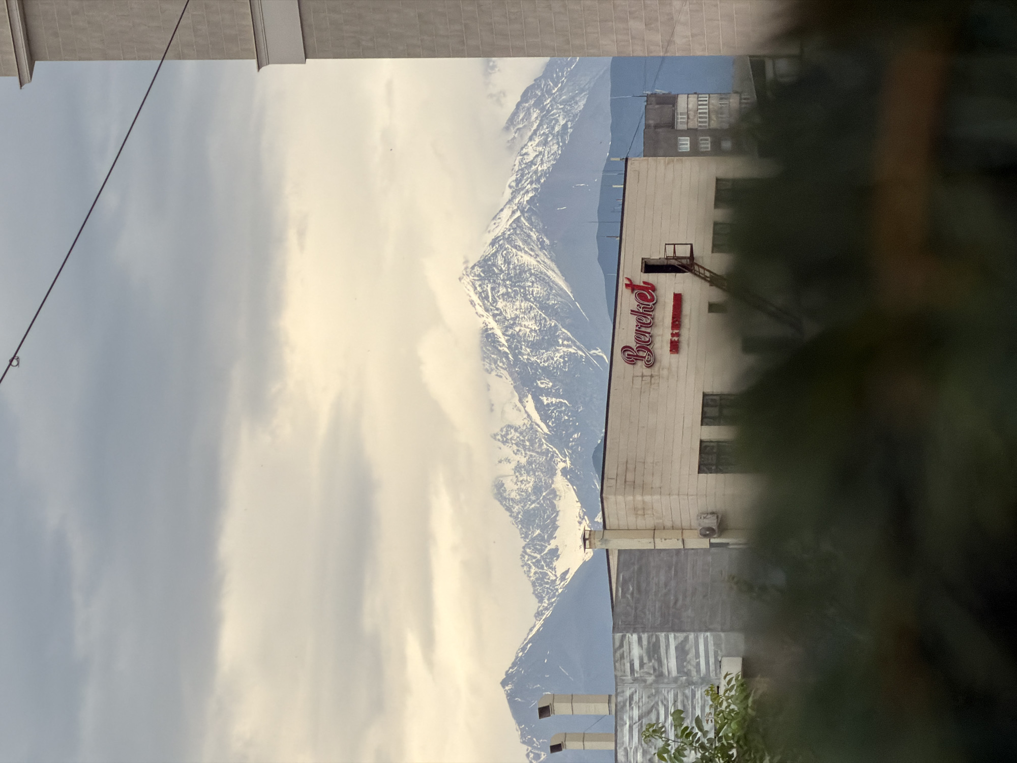 In Bishkek City, Kyrgyzstan, a low-angle shot reveals snow-capped mountains in the distance under a cloudy sky. In the foreground, a building with a sign that reads "Bireket CAFE & RESTAURANT" in red script is visible. A fire escape leads up to an open door on the building. To the left of this building is a gray structure with silver siding, and to the right, a portion of a light-colored building with decorative trim is seen. Further to the right, a dark gray apartment building with multiple windows stands against the skyline. The foreground is partially obscured by dark green foliage.