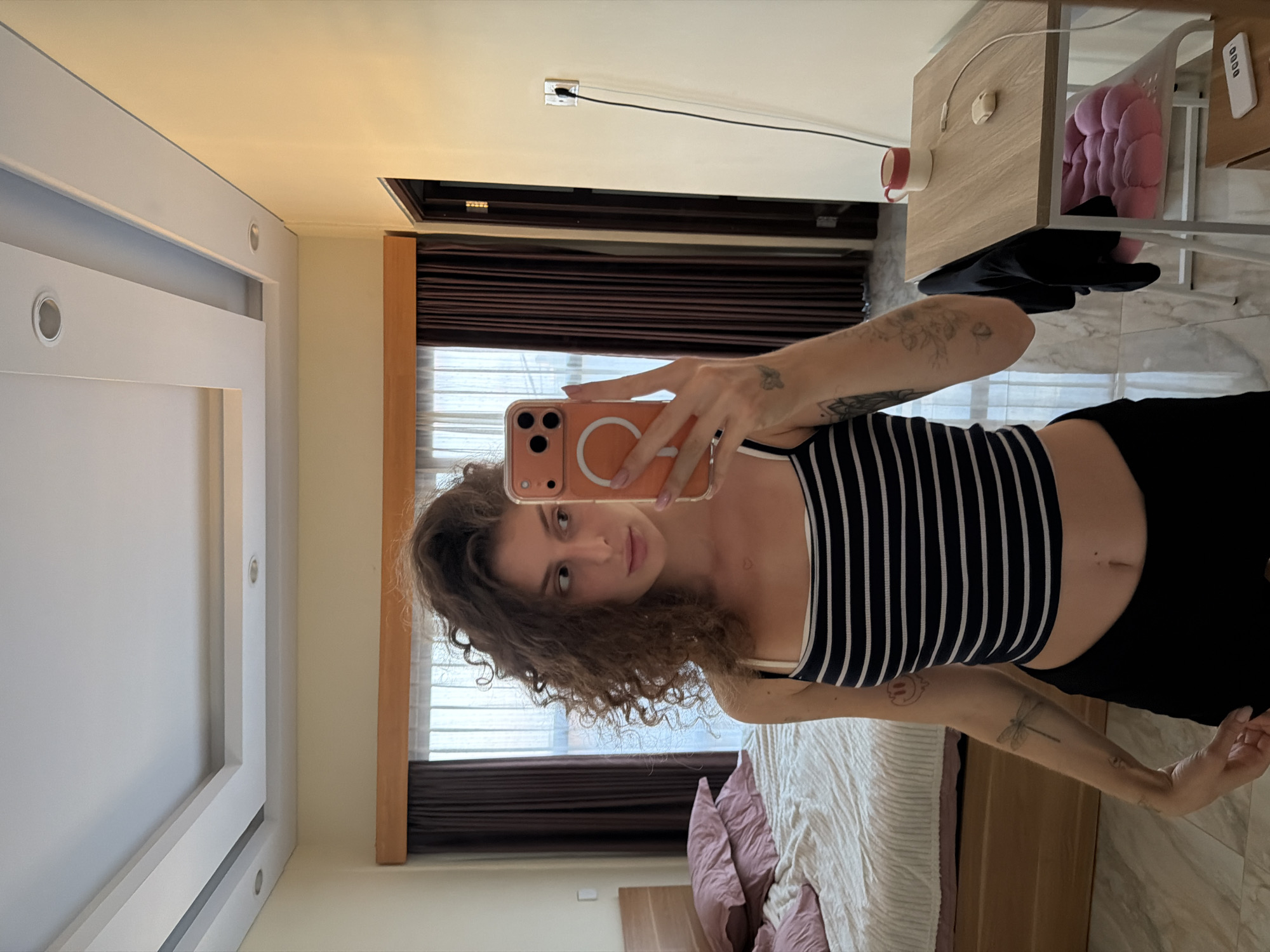 A young woman with curly brown hair stands in a bedroom, taking a selfie in a mirror. She is wearing a black and white striped crop top and black shorts. Her left arm has several tattoos, including a dragonfly. A bed with pink pillows is visible to her left, and a small wooden desk with a mug, a phone charger, and a pink cushion on a chair is to her right. The room has light-colored walls and a white ceiling with recessed lighting. The overall setting appears to be a hotel room in North Kuta, Indonesia, as suggested by the presence of typical hotel furnishings.