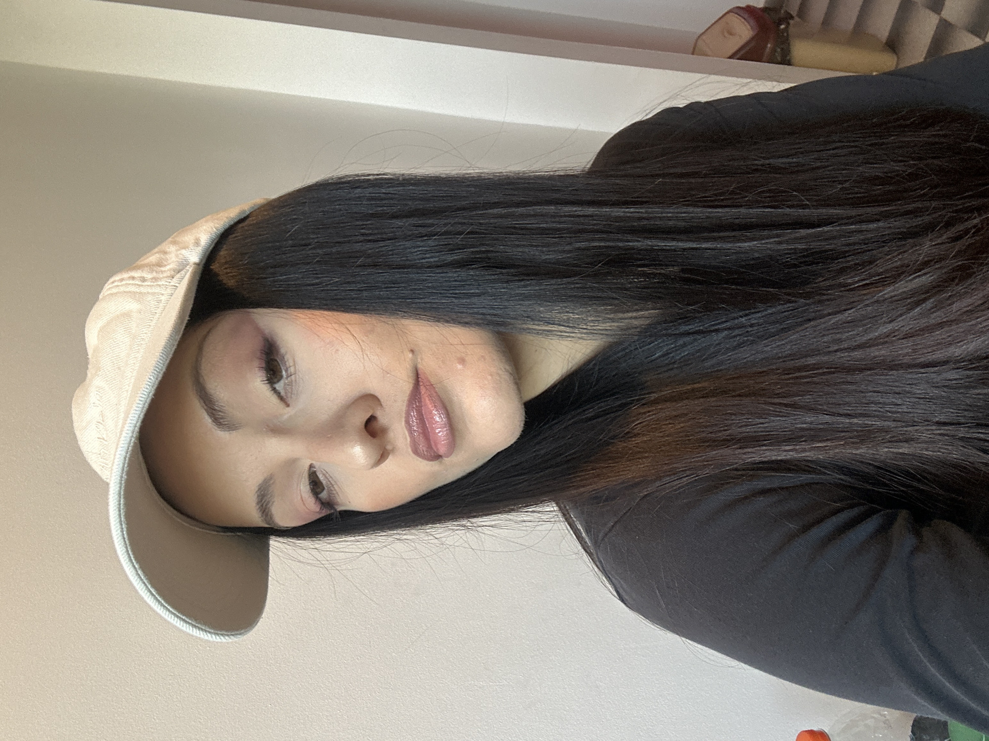 A young woman with long, dark hair is wearing a light-colored baseball cap. She is facing the camera with her head tilted to the side. She is wearing makeup. The background is a plain, light-colored wall. No location information from Burgas, Bulgaria is discernible.