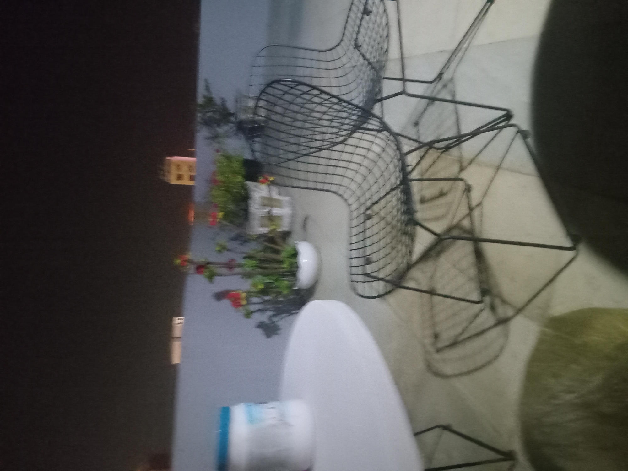 The image depicts an outdoor setting, likely a balcony or patio, in Đà Nẵng, Vietnam. The scene features several black metal wire chairs, a common style for outdoor furniture, arranged around a white, oval-shaped table. To the left of the table, a potted plant with green foliage and small red and yellow flowers sits on a ledge. In the background, a blurry building with illuminated windows can be seen against a dark night sky, suggesting the photo was taken at night. The overall atmosphere is tranquil, with no people visible. The lighting suggests artificial illumination from the table area and the distant building.