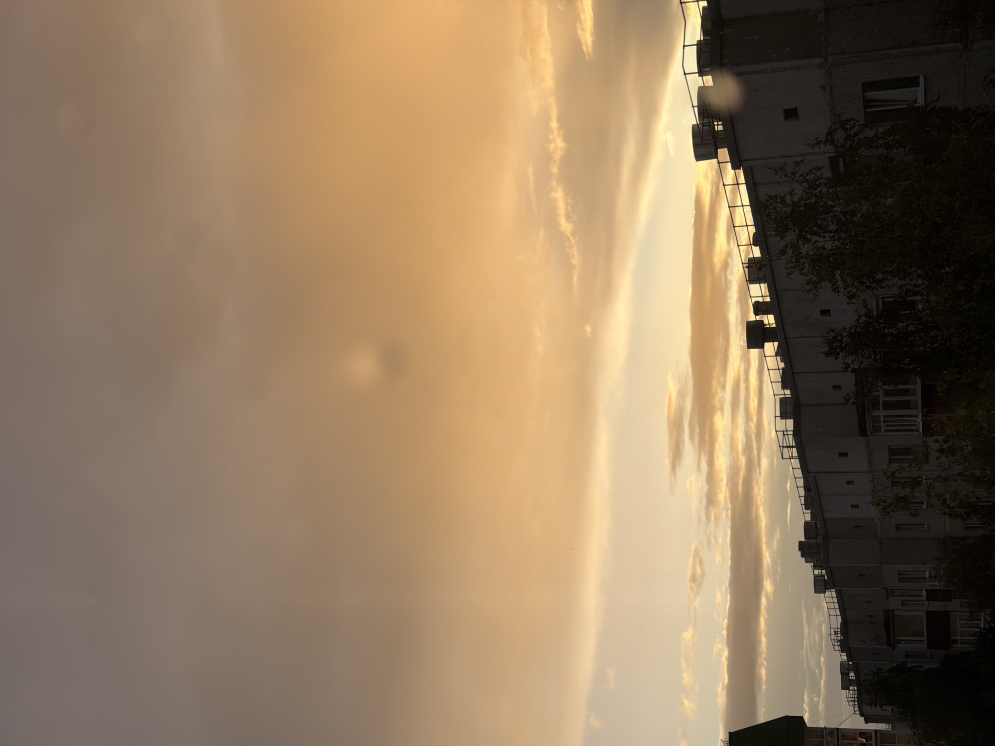 An outdoor, eye-level, medium shot captures a sunset over an apartment building in Surgut, Russia. The sky is filled with a mix of orange and gray clouds. A faint, circular distortion is visible in the upper center of the sky. Below the sky, the upper portion of a large, gray apartment building is visible, with a railing along its roof. Several cylindrical structures, possibly vents or chimneys, protrude from the roof. Trees are partially obscuring the lower sections of the building. To the left, a portion of another building with a dark green roof is visible. The overall scene is dimly lit due to the time of day.