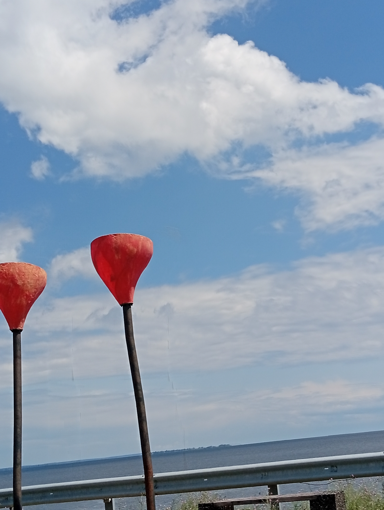 Two red, funnel-shaped buoys stand tall against a backdrop of a cloudy blue sky and a calm body of water. The buoys are mounted on dark, slender poles, with the buoy on the left appearing slightly smaller and more weathered than the one on the right. A metal guardrail runs horizontally across the lower portion of the image, separating the buoys from the water. Beyond the guardrail, the water stretches to the horizon, meeting a distant, hazy shoreline. The sky is filled with fluffy white clouds, some large and billowy, others wispy and scattered. The overall scene suggests an outdoor setting, possibly near a waterfront or along a coastal road.