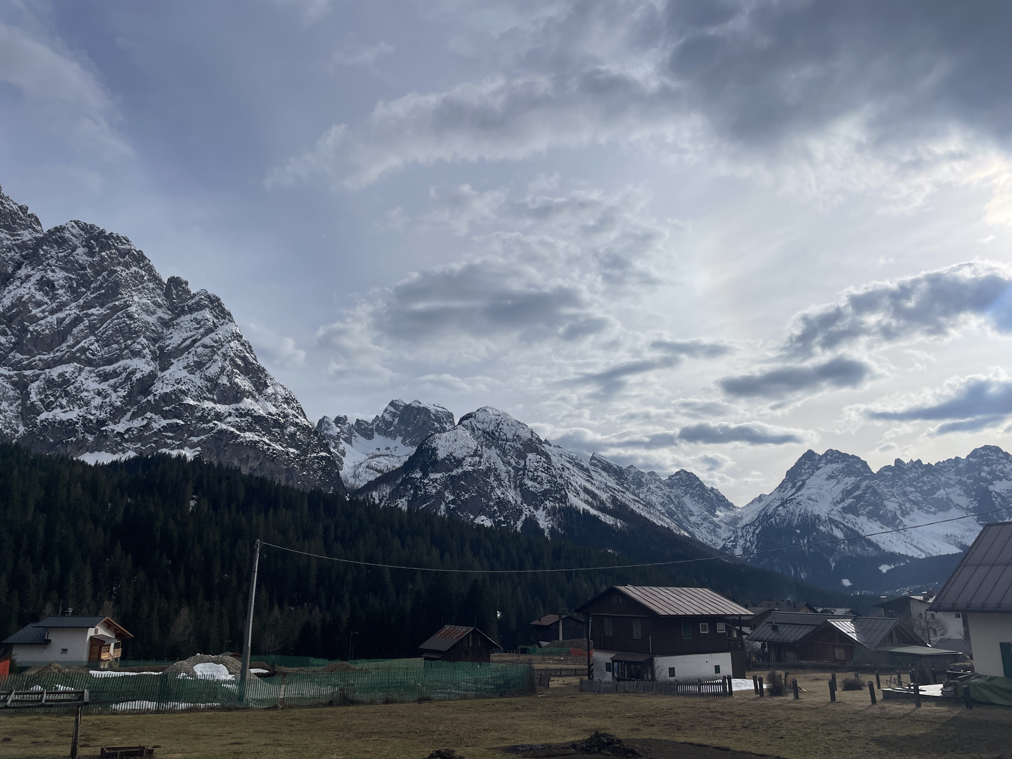 The image depicts a mountainous landscape in Sappada, Italy. Snow-capped mountains dominate the background, with a dense forest covering the lower slopes. In the foreground, there is a field with several wooden houses and buildings scattered across it. The sky is overcast with grey clouds, and the light appears to be diffused. There are no people visible in the scene.